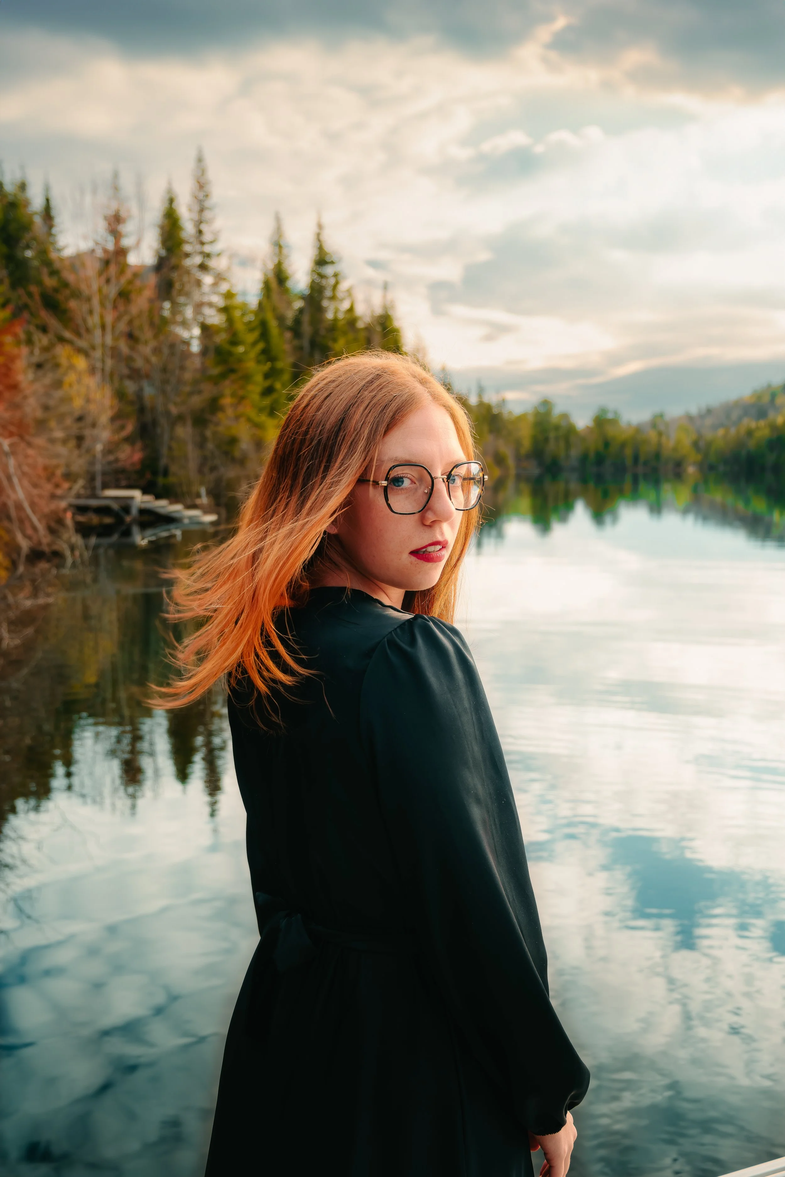 Jeune femme aux cheveux roux portant des lunettes, regardant par-dessus son épaule près d'un lac, avec des arbres et un ciel nuageux en arrière-plan.