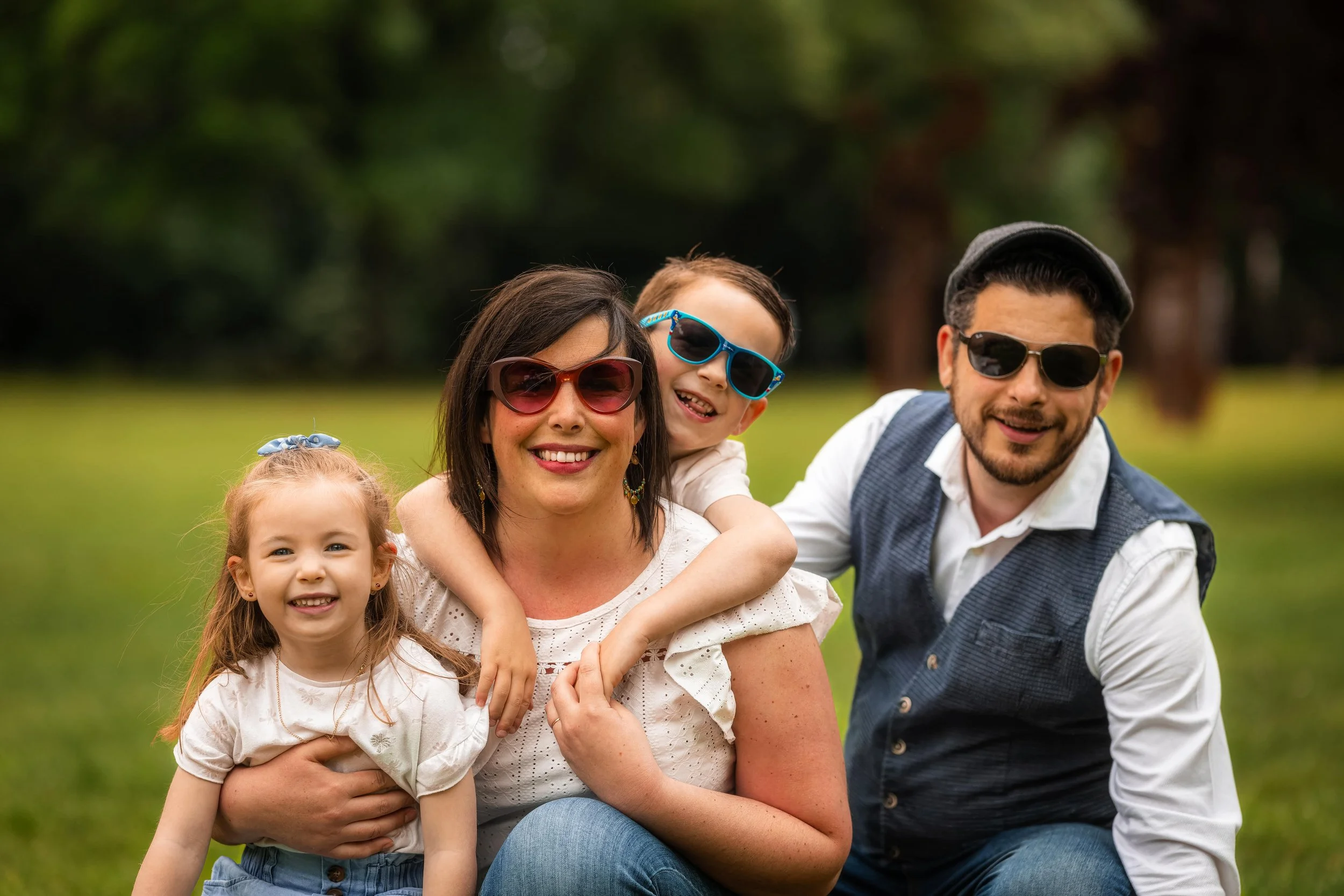 Famille de quatre personnes souriantes, posant dans un parc, portant des lunettes de soleil, avec un fond vert naturel.