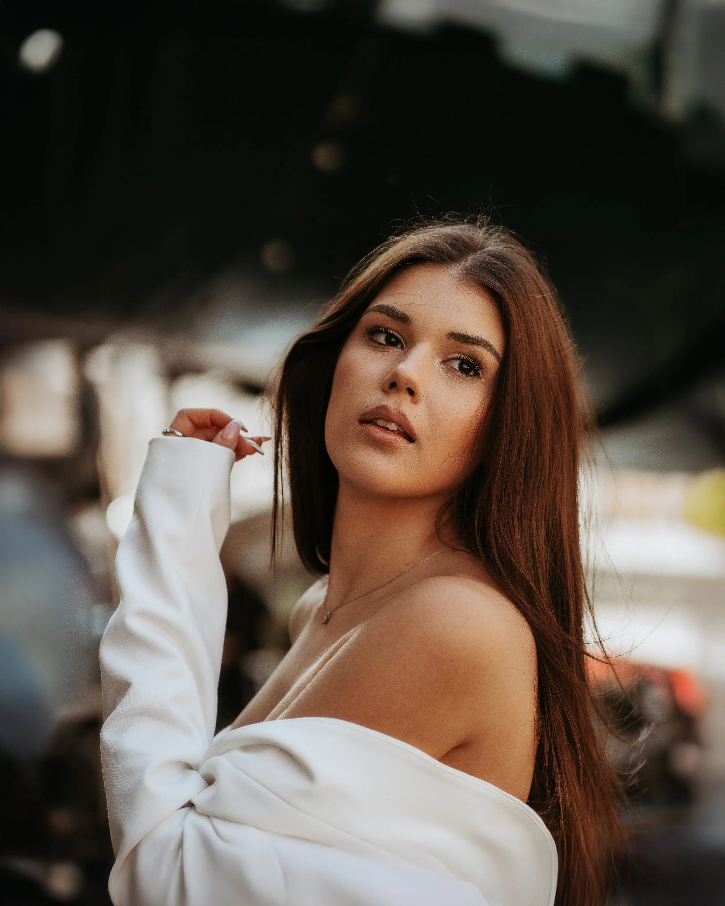Jeune femme à la peau claire, cheveux bruns, portant une robe blanche avec une épaule dénudée, posant à l'extérieur avec un fond flou de structures urbaines.