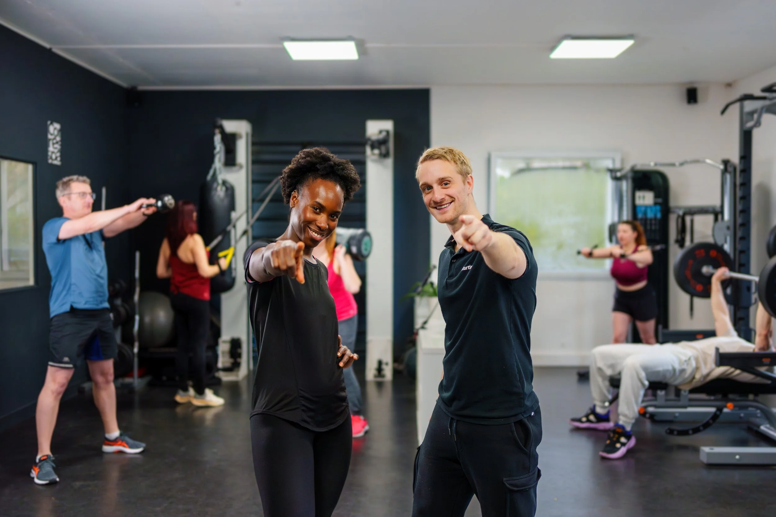 Deux entraîneurs souriants pointant vers la caméra, dans une salle de sport avec plusieurs personnes qui s'entraînent en arrière-plan.