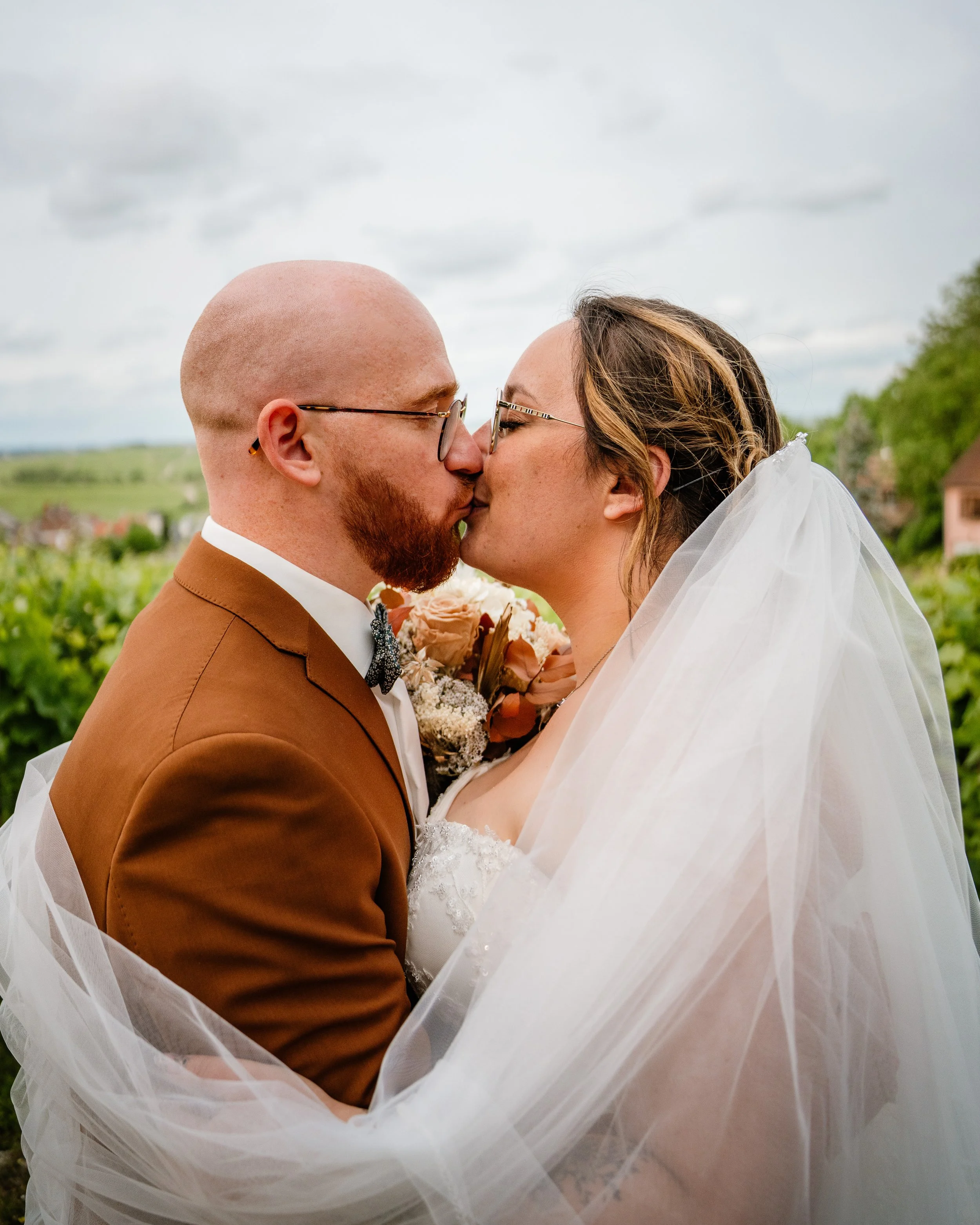 Un couple marié s'embrassant en plein air, arrière-plan de champs et de maisons.