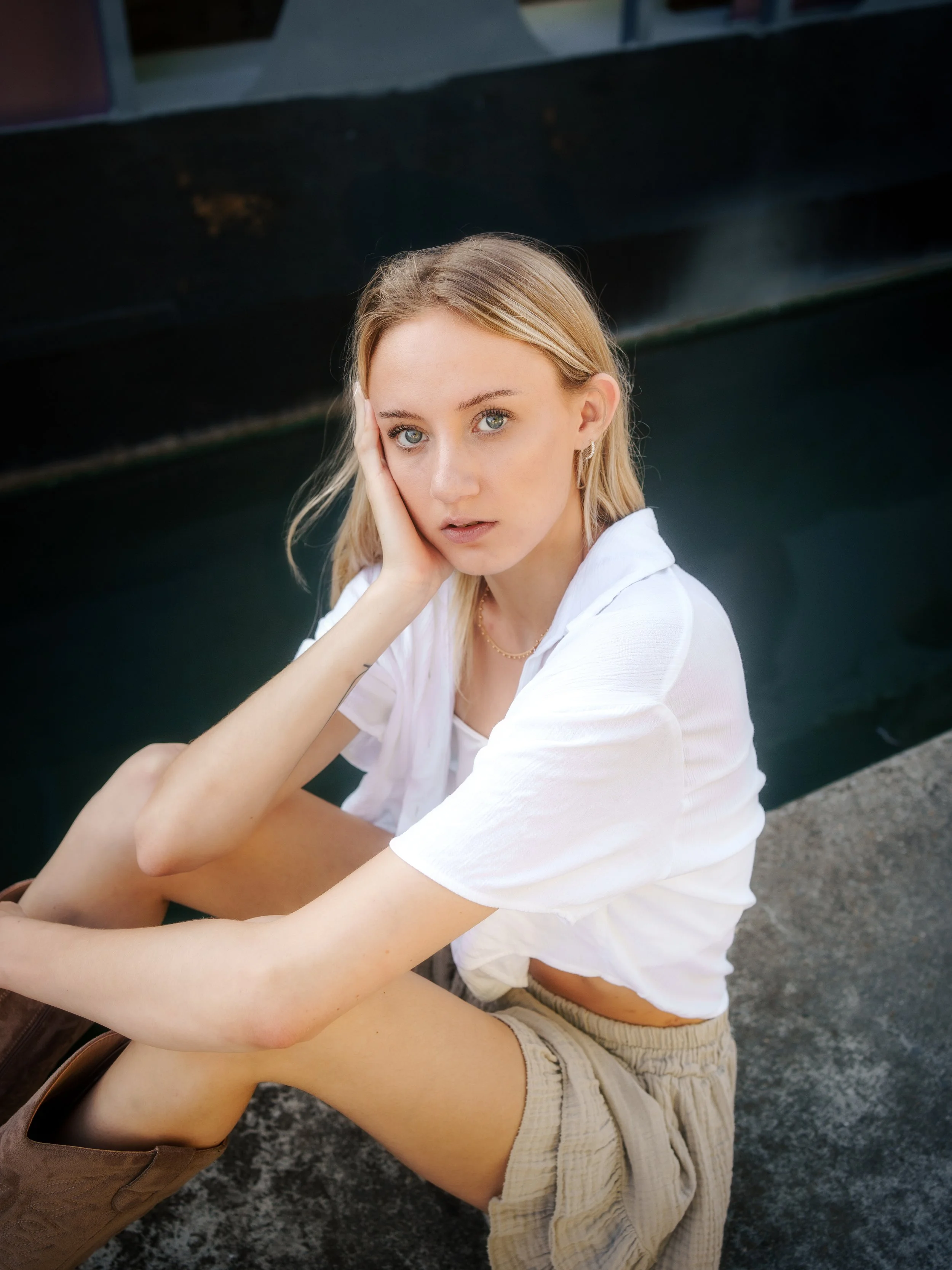Jeune femme assise sur le sol près d'un bassin d'eau, portant un t-shirt blanc, un short beige et des bottes marron, regardant vers l'appareil photo avec une expression pensif.