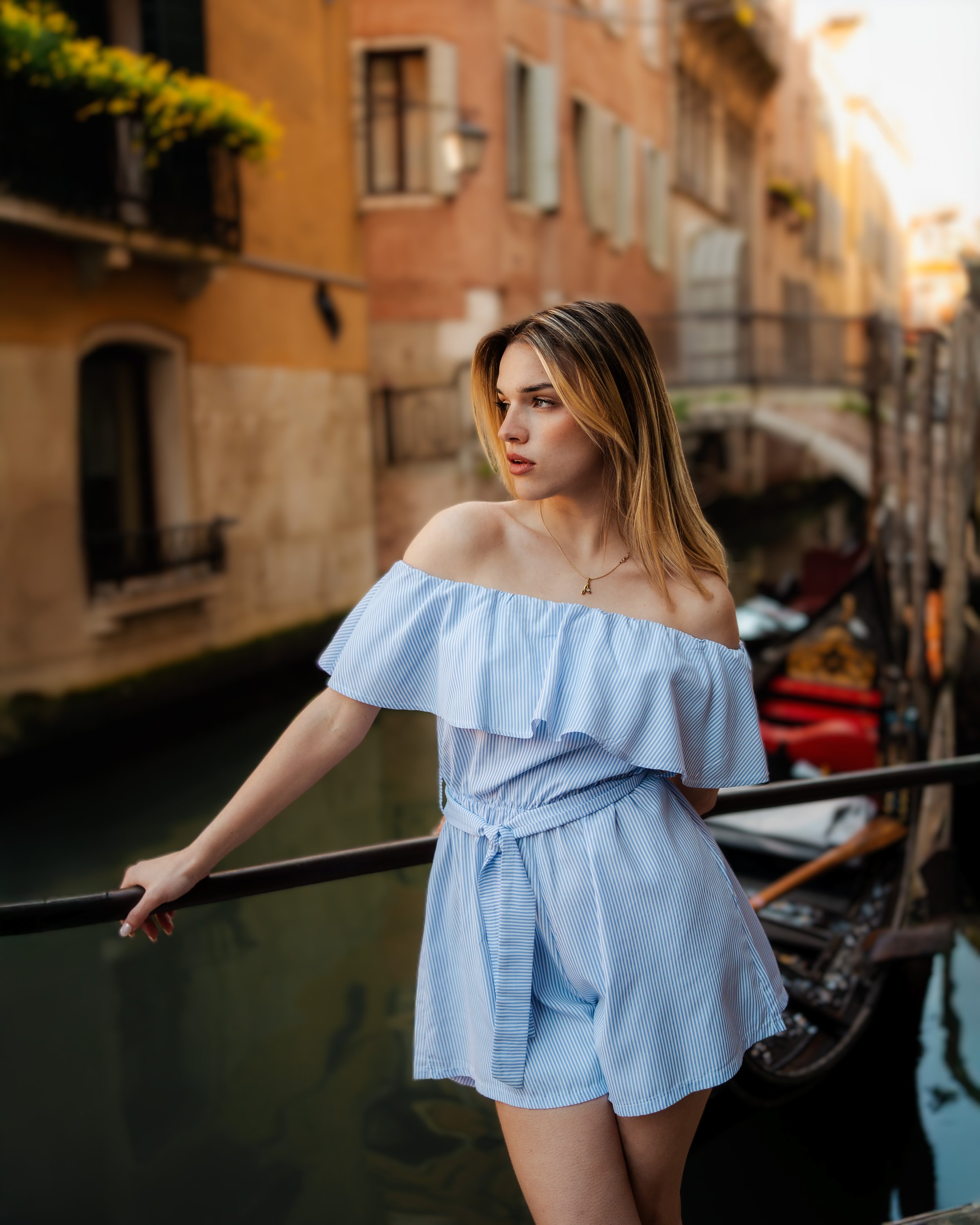 Jeune femme portant une robe à rayures bleues et blanches, vêtue d'une robe d'été à épaules dénudées, se tenant à une barrière le long d'un canal italien, avec des bâtiments colorés en arrière-plan.