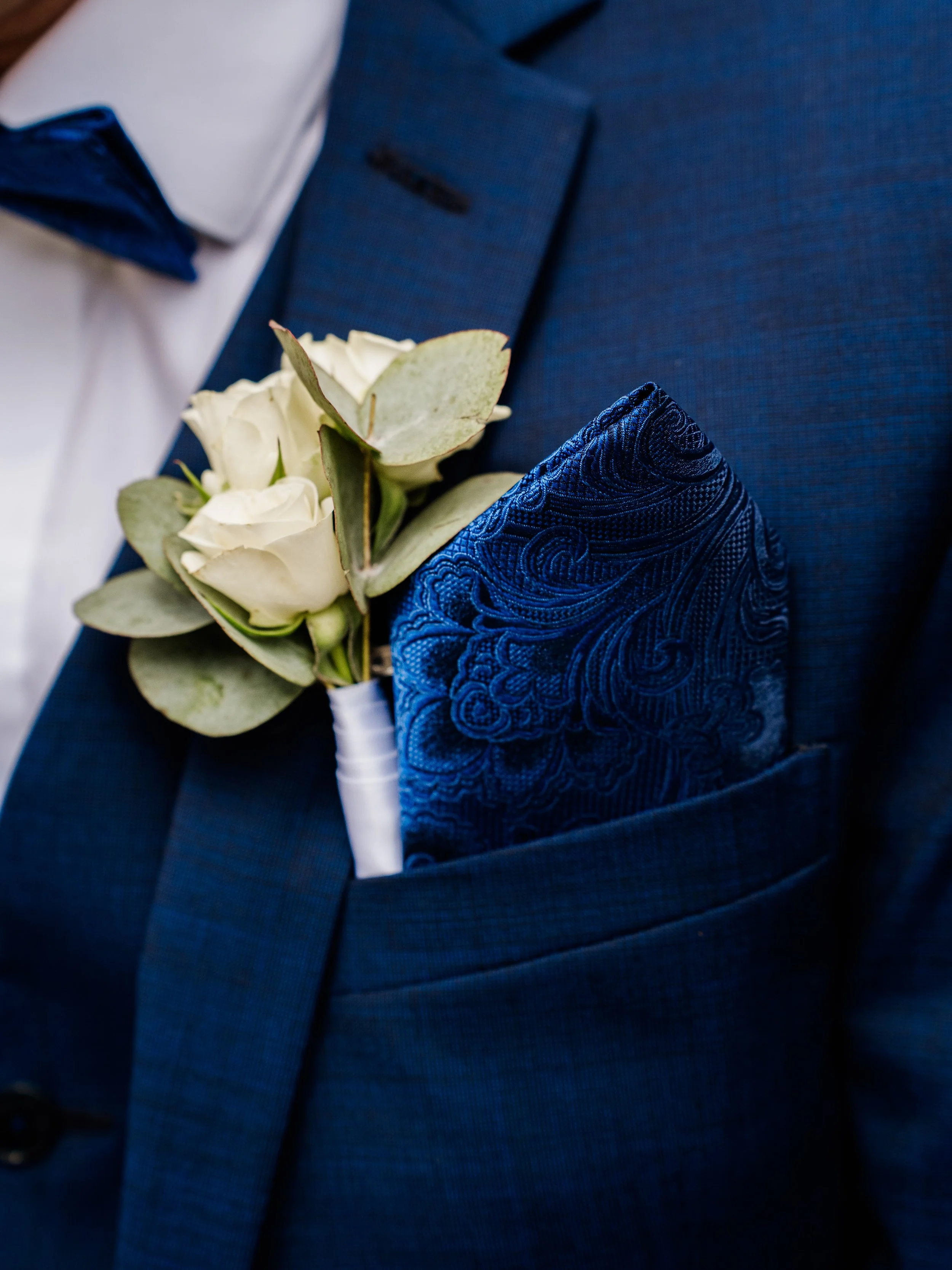 Boutonnière avec des fleurs blanches et des feuilles, dans la poche de la veste bleue d'un homme en costume.