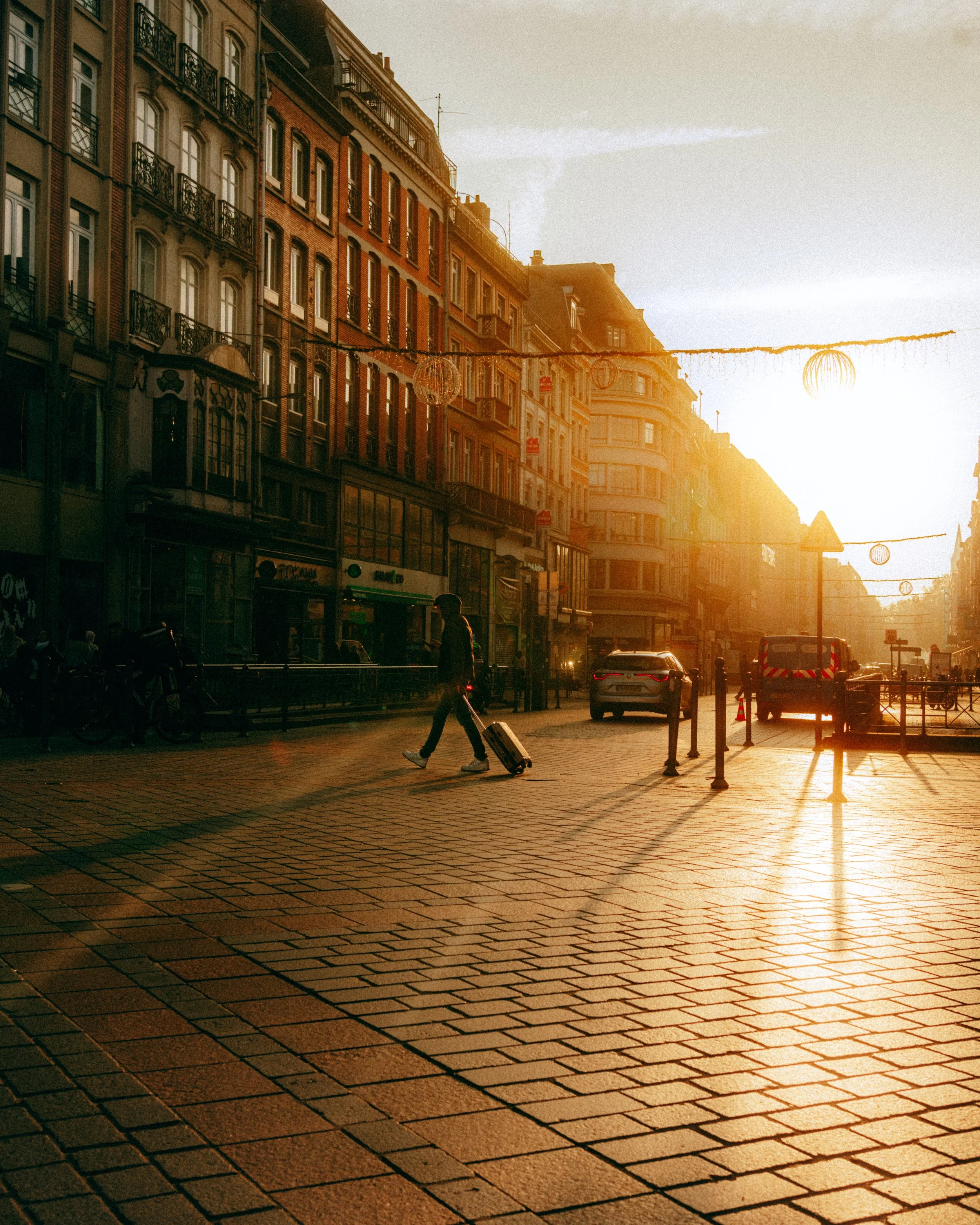 Une rue pavée d'une ville européenne avec des bâtiments anciens en brique, des lampadaires et des décorations suspendues, au coucher du soleil, avec une personne marchant avec une valise.