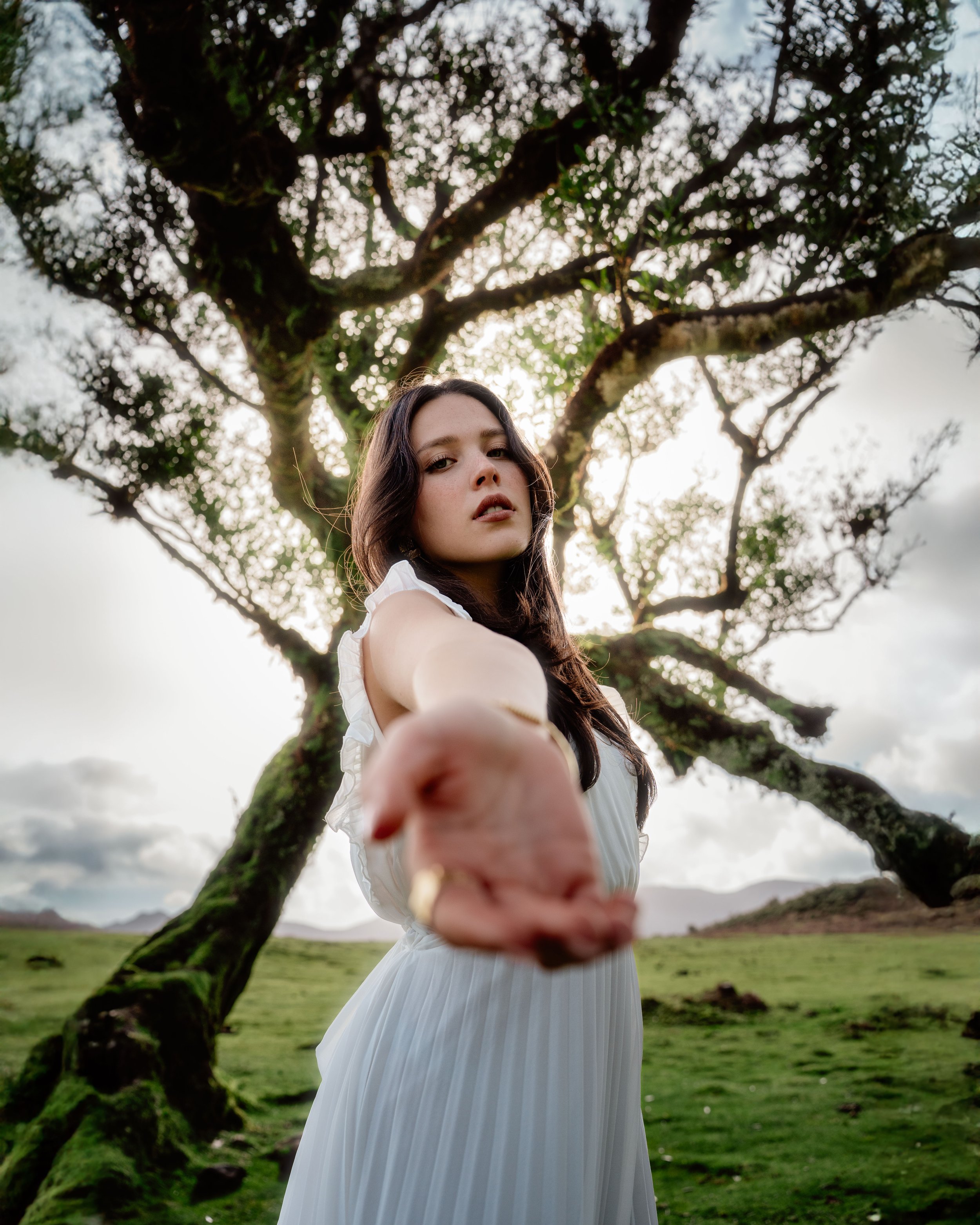 Jeune femme en robe blanche, debout dans un paysage champêtre avec un arbre en arrière-plan, elle tend la main vers la caméra.