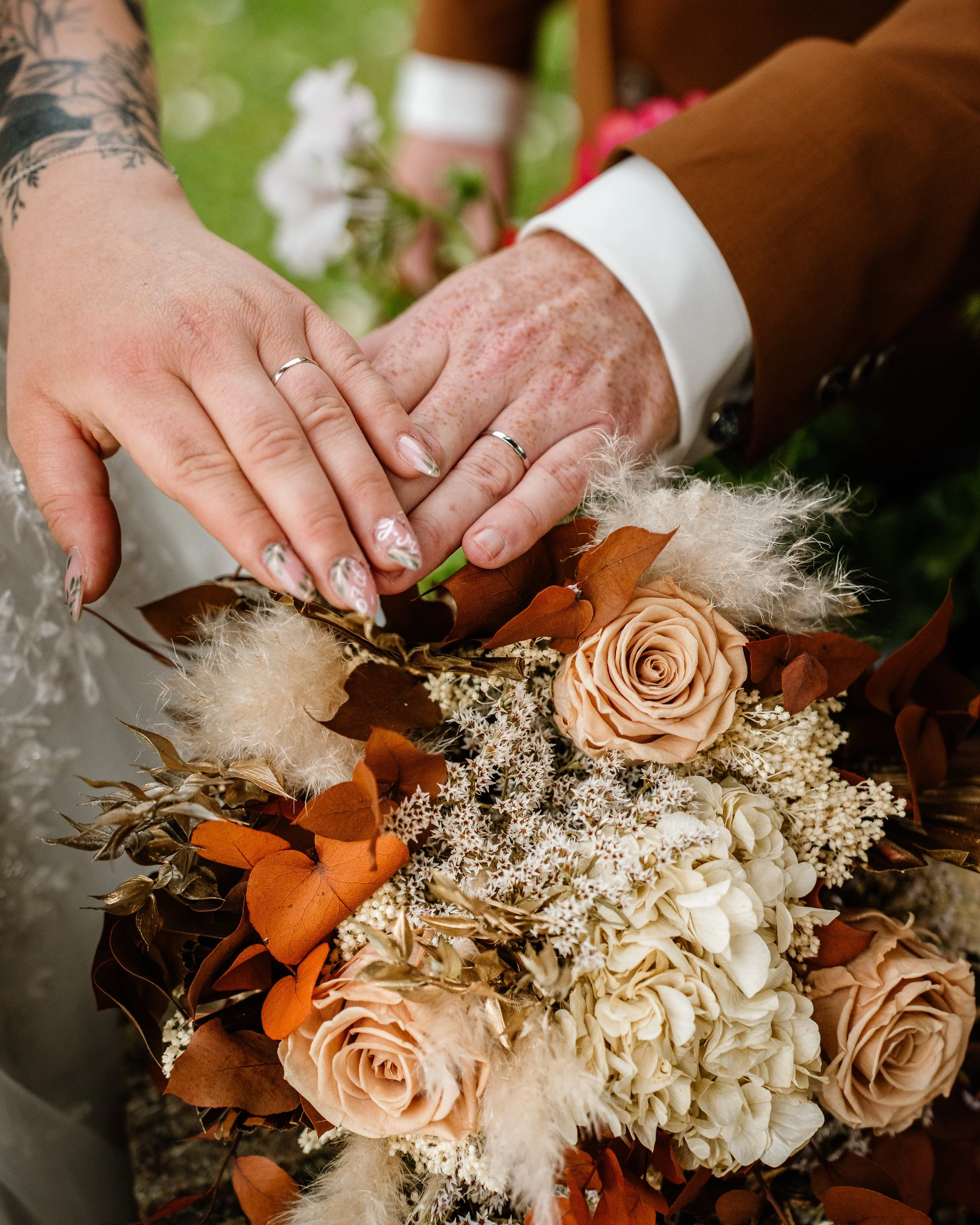 Les mains de deux personnes portant des alliances reposent sur un bouquet de fleurs avec des roses beige, des feuilles marron et des éléments d'ornement blanc