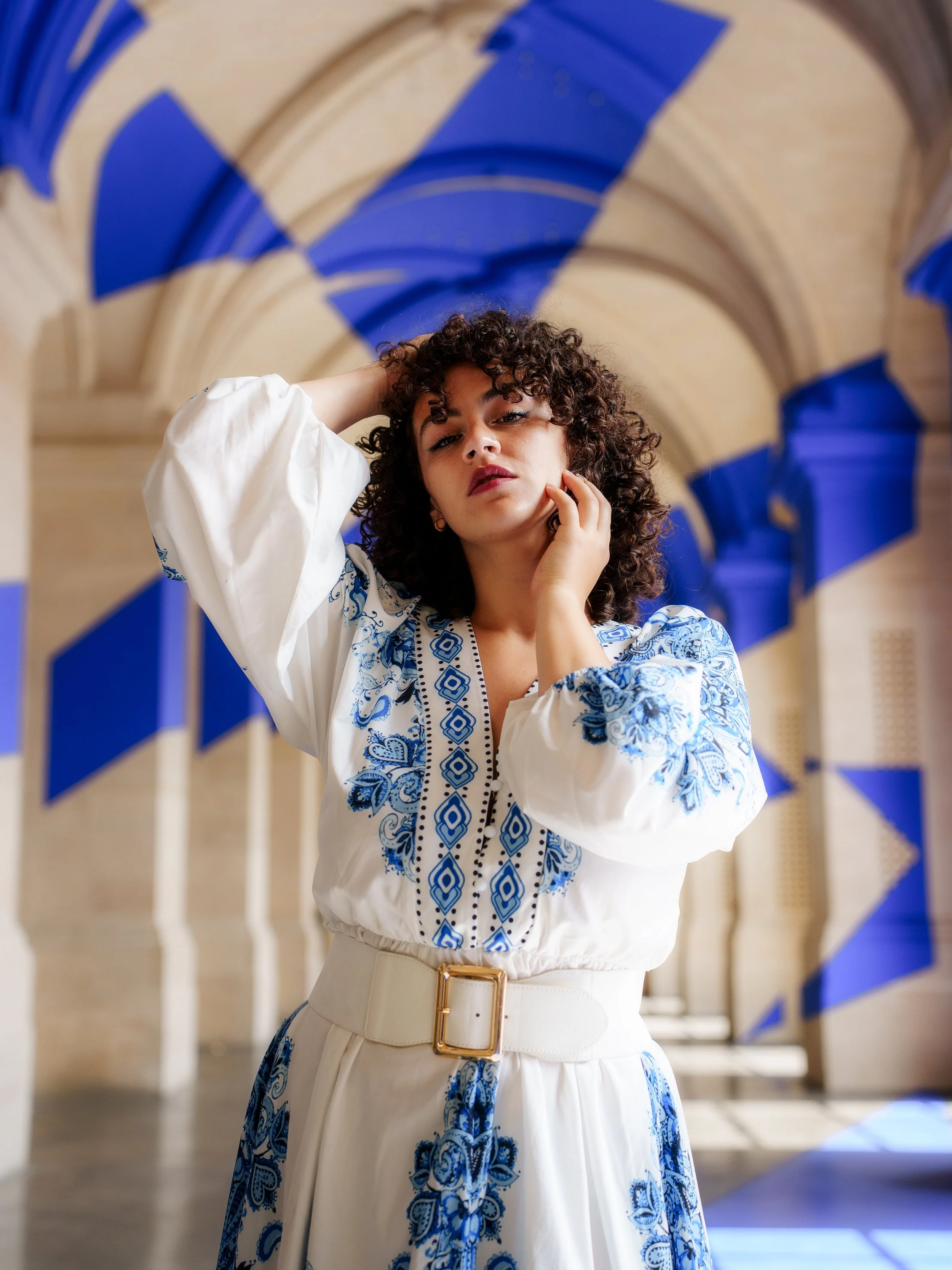 Une femme avec des cheveux bouclés et un maquillage subtil, portant une robe blanche avec des broderies bleues, dans un espace archétypal avec des arches en pierre et des décorations bleues.