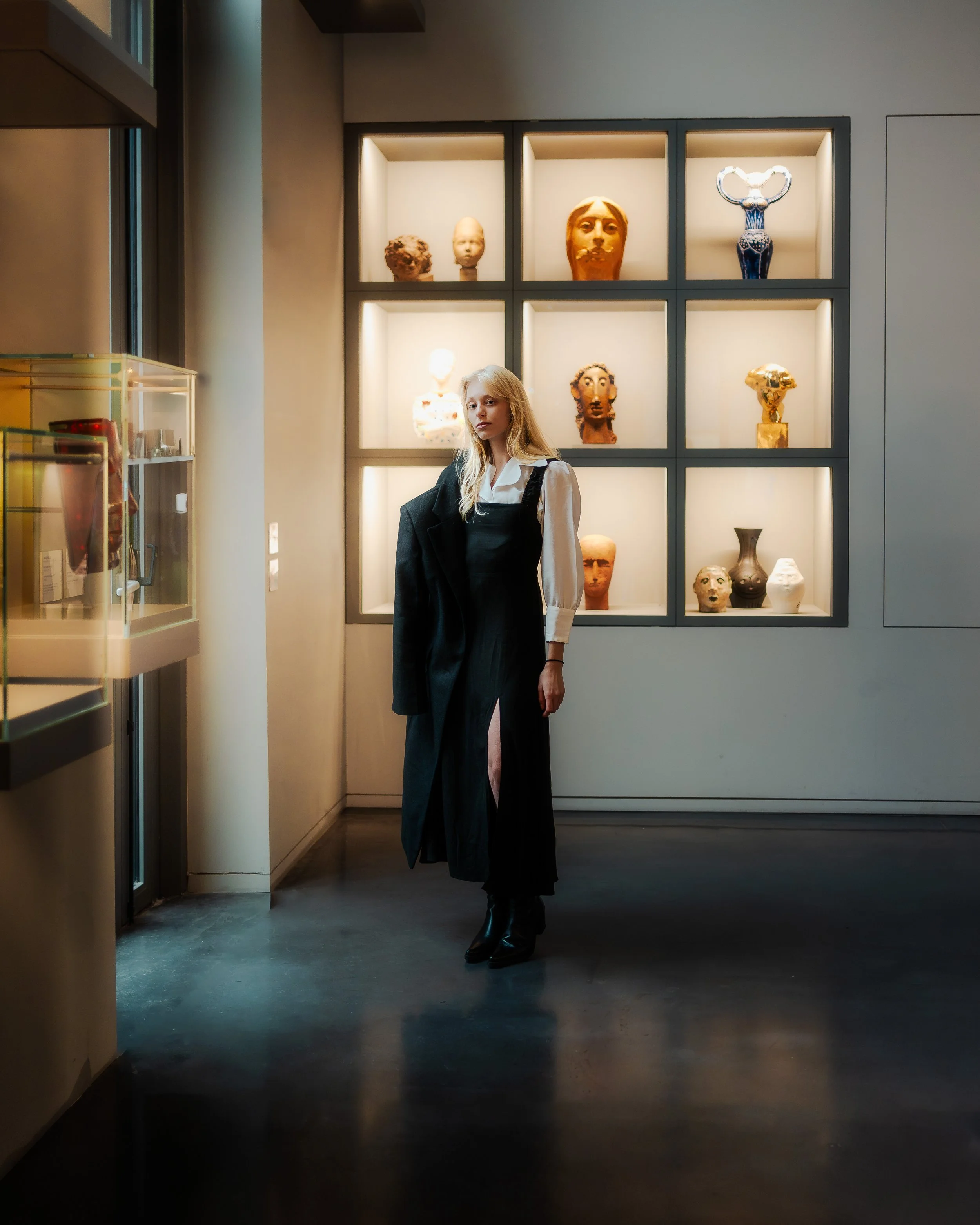 Jeune femme dans un musée, debout devant une vitrine contenant des objets, portant une robe noire avec une veste noire suspendue à son bras, et un fond de murs avec des sculptures exposées dans des niches éclairées.