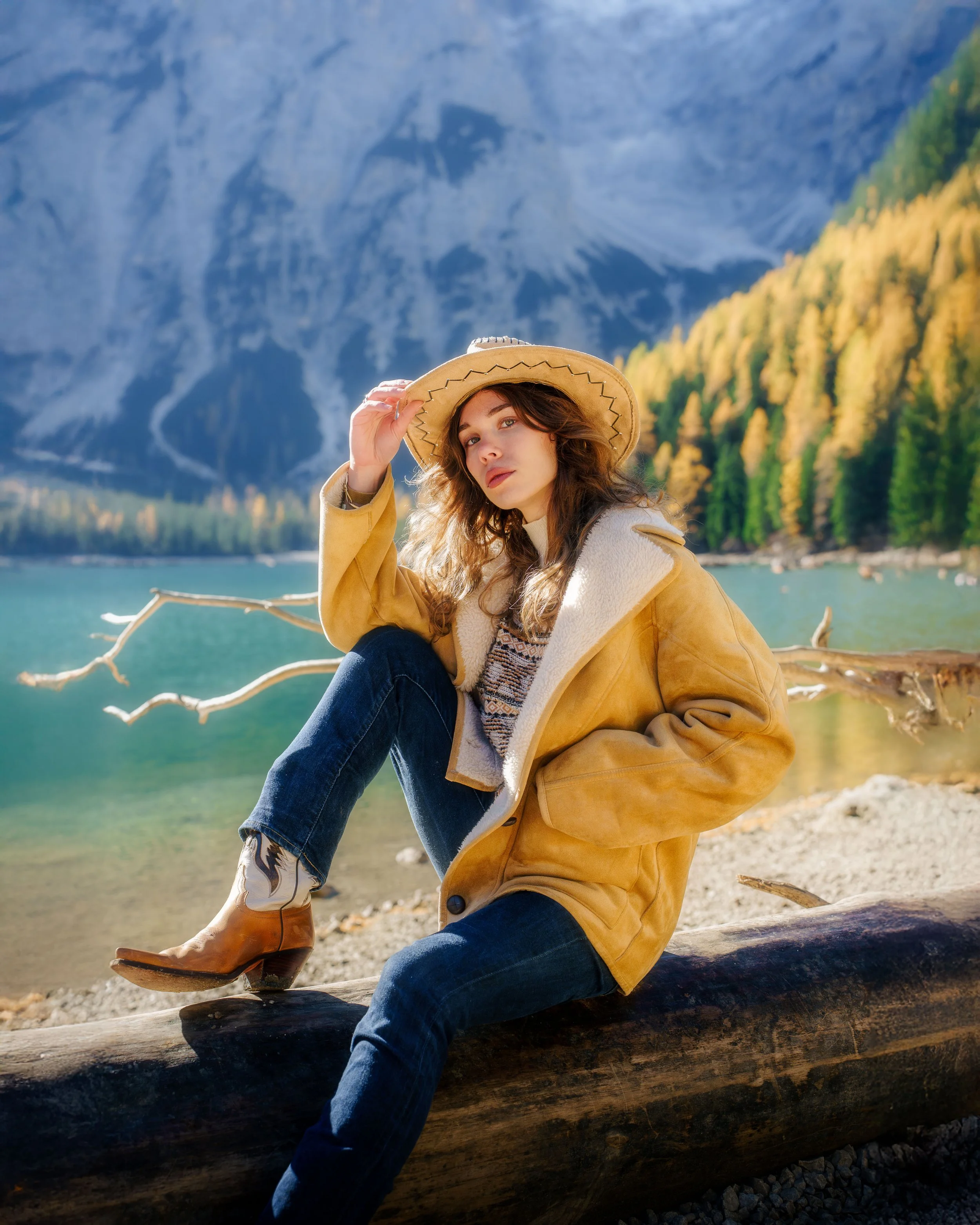Une jeune femme en veste en daim et chapeau de paille, assise sur une branche d'arbre au bord d'un lac de montagne entouré d'arbres et de montagnes, sous un ciel ensoleillé.