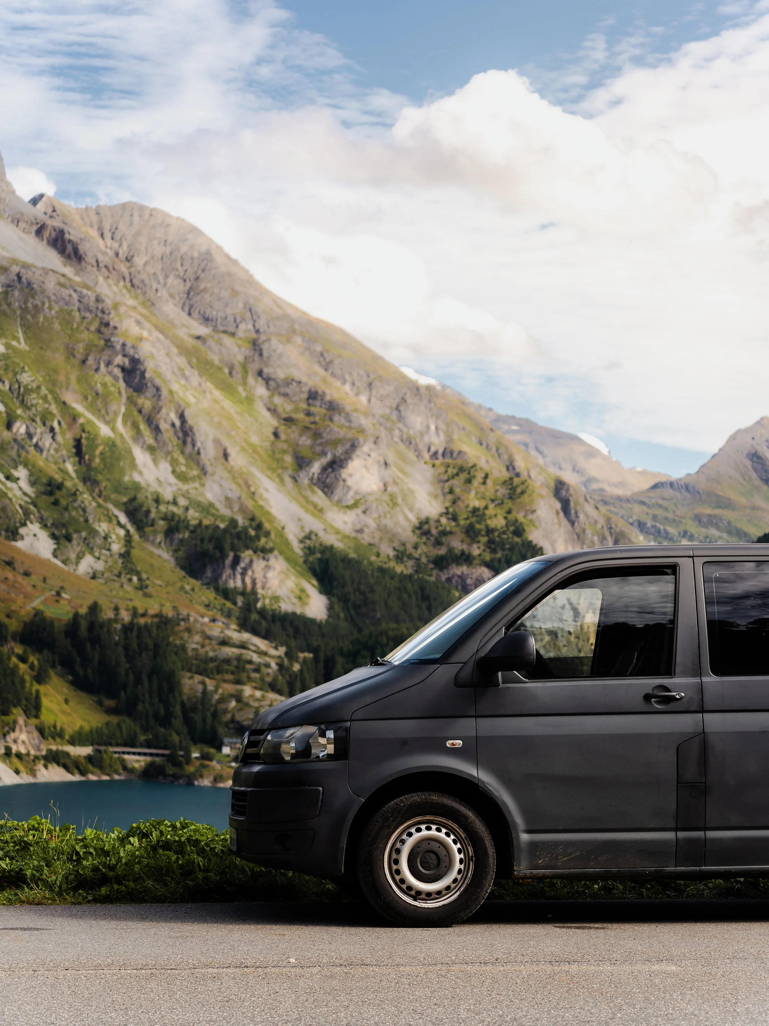 Voiture noire stationnée sur une route au bord d'un lac entouré de montagnes verdoyantes sous un ciel partiellement nuageux.