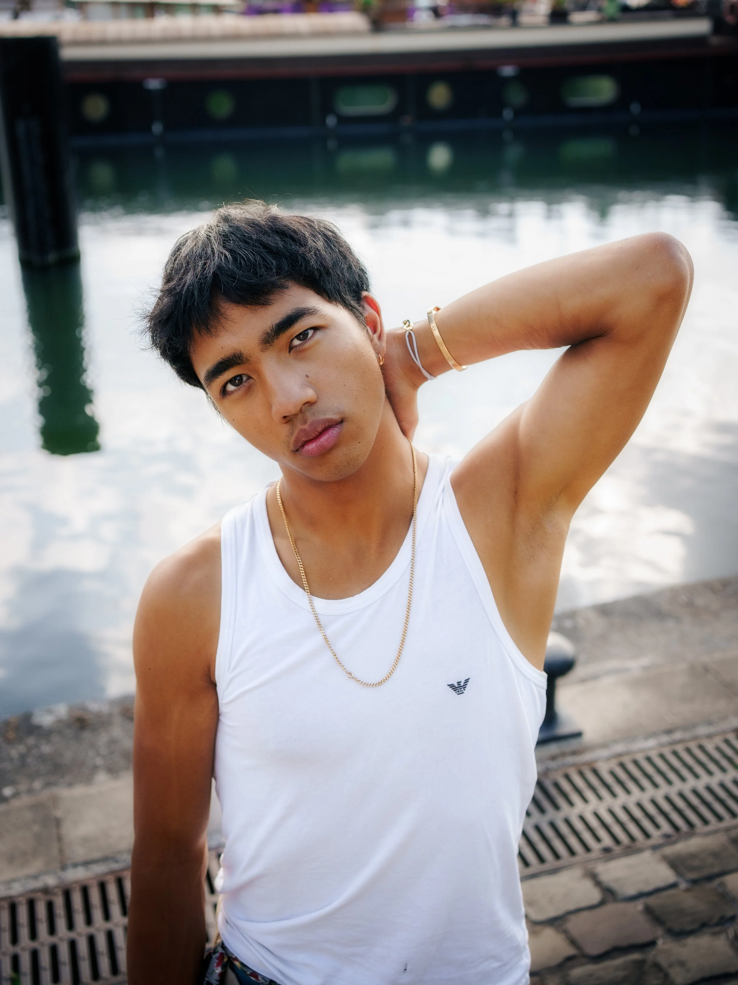 Jeune homme posant près d'un plan d'eau, portant un débardeur blanc, des bijoux en or, avec fond d'eau et pont en arrière-plan.