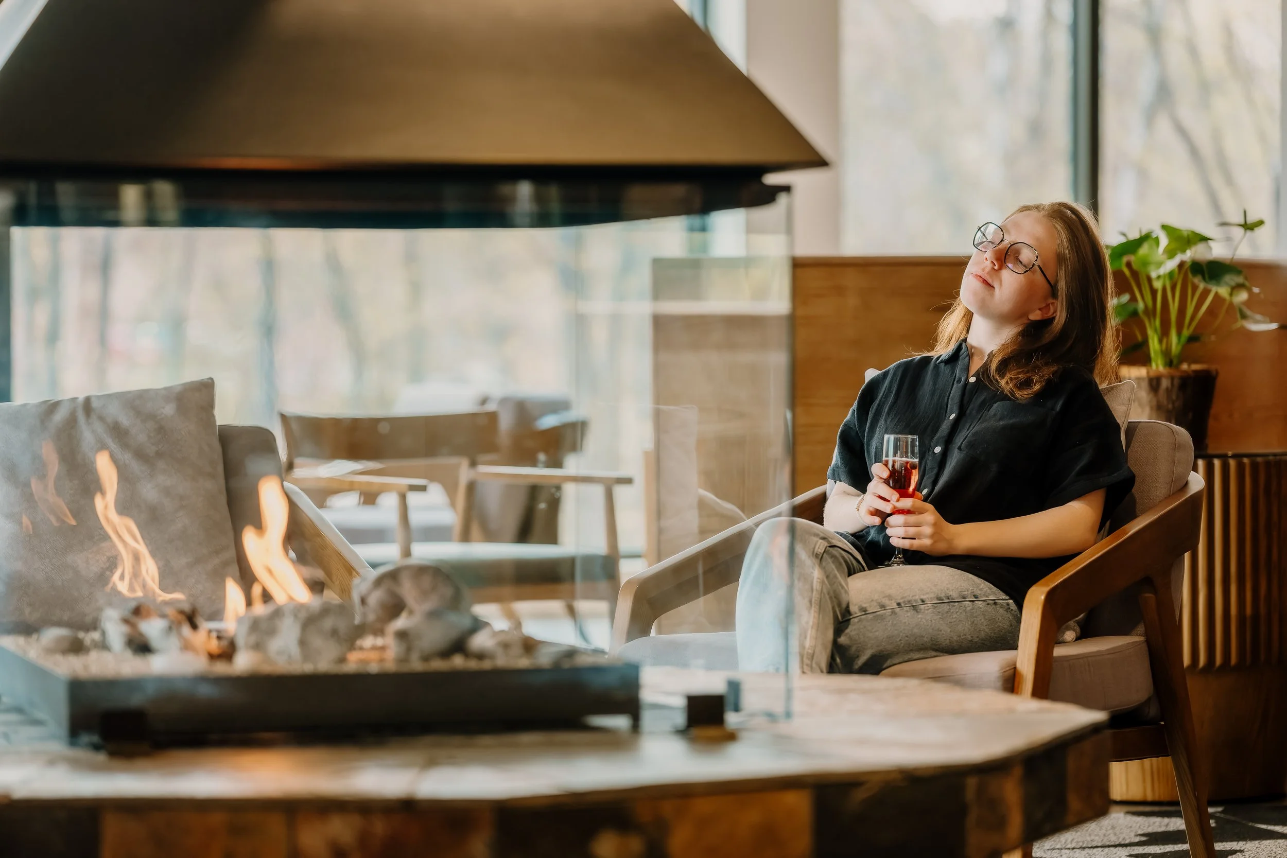 Une femme assise dans un salon près d'une cheminée, tenant un verre de boisson rouge, avec une fenêtre en arrière-plan laissant entrer de la lumière naturelle.