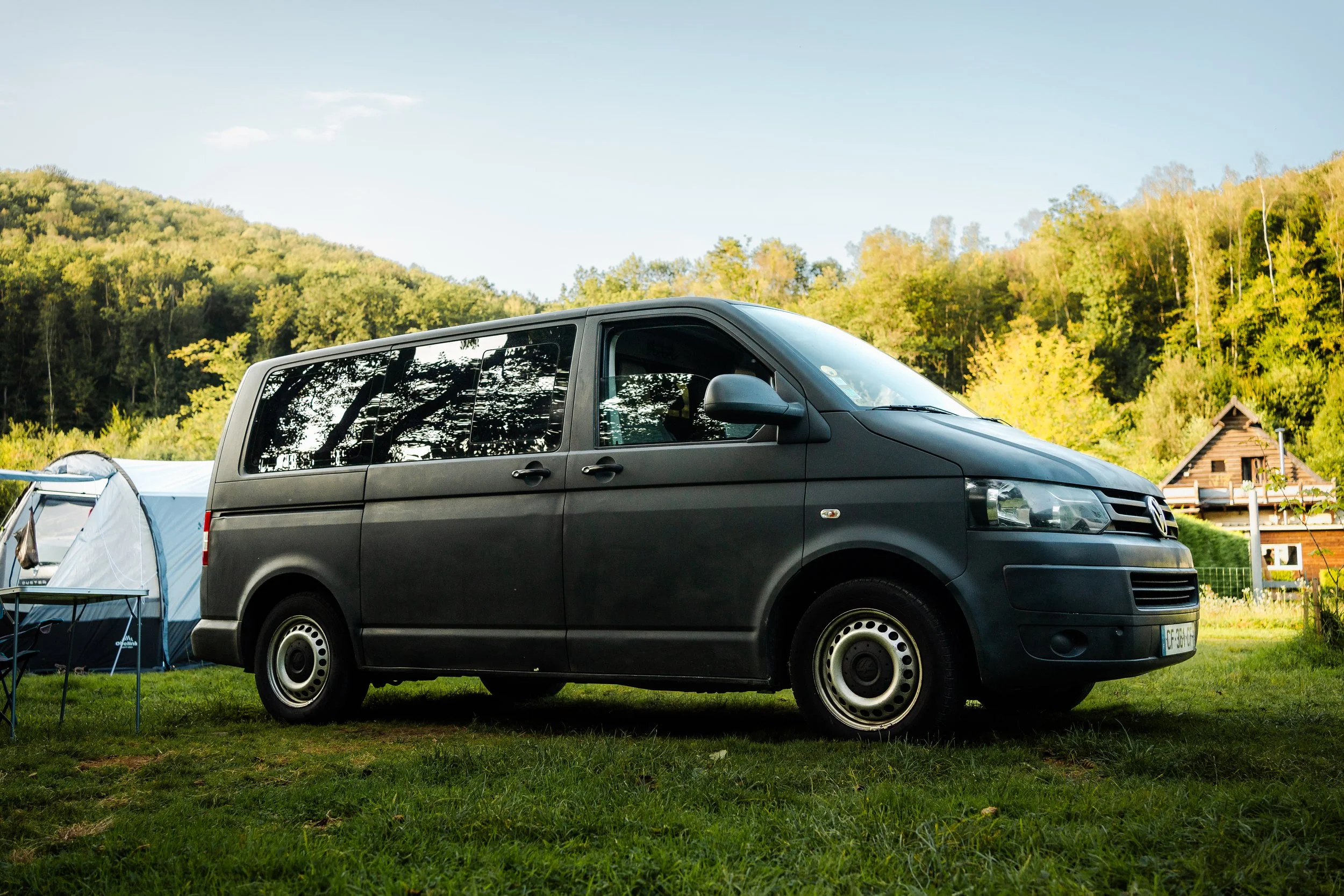 Véhicule utilitaire noir stationné sur un terrain verdoyant avec tentes et une maison en bois en arrière-plan, dans un environnement naturel avec des collines et des arbres.