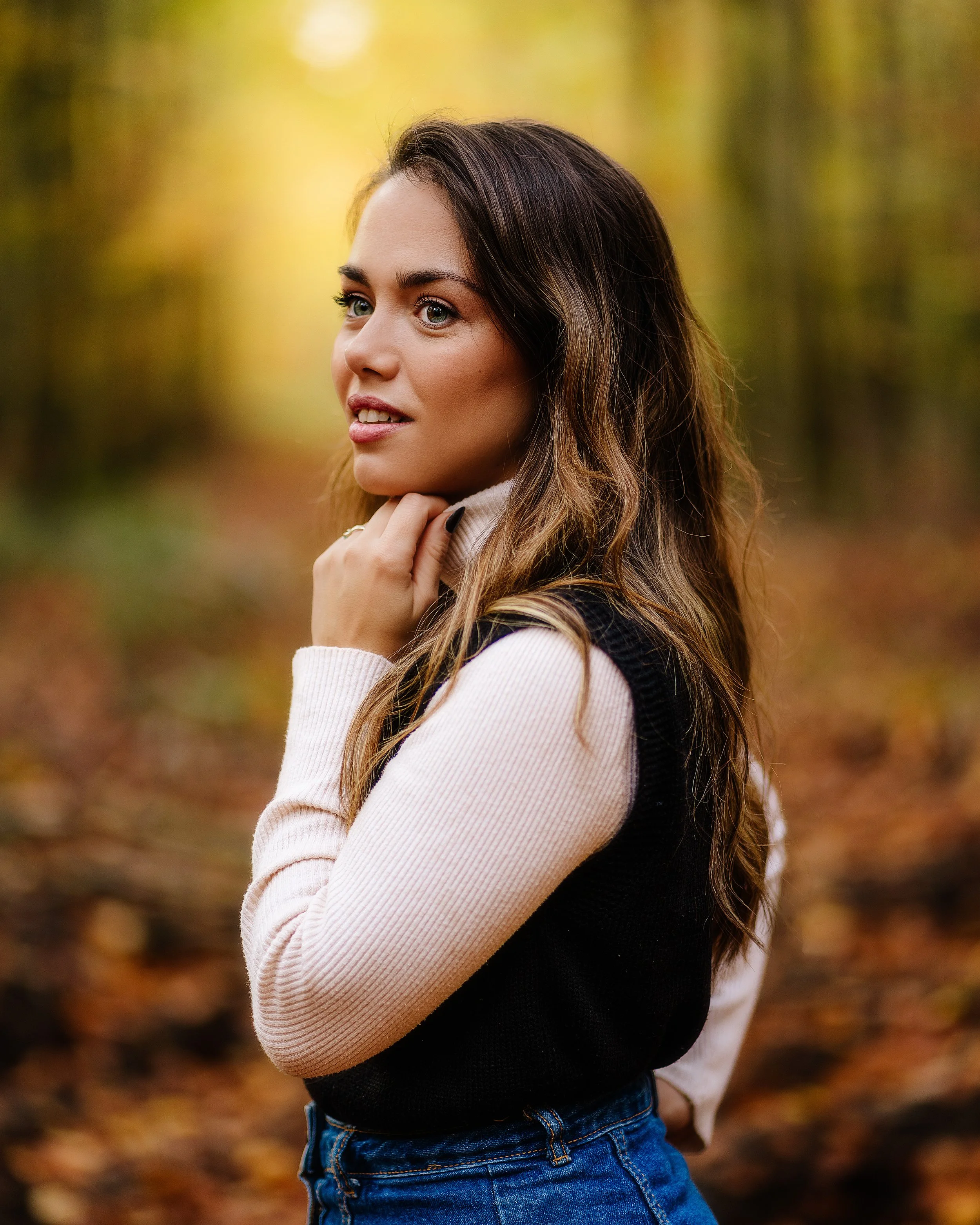 Jeune femme posant dans une forêt automnale, portant un pull rayé beige et un gilet noir, avec des cheveux longs et ondulés, regardant à gauche.