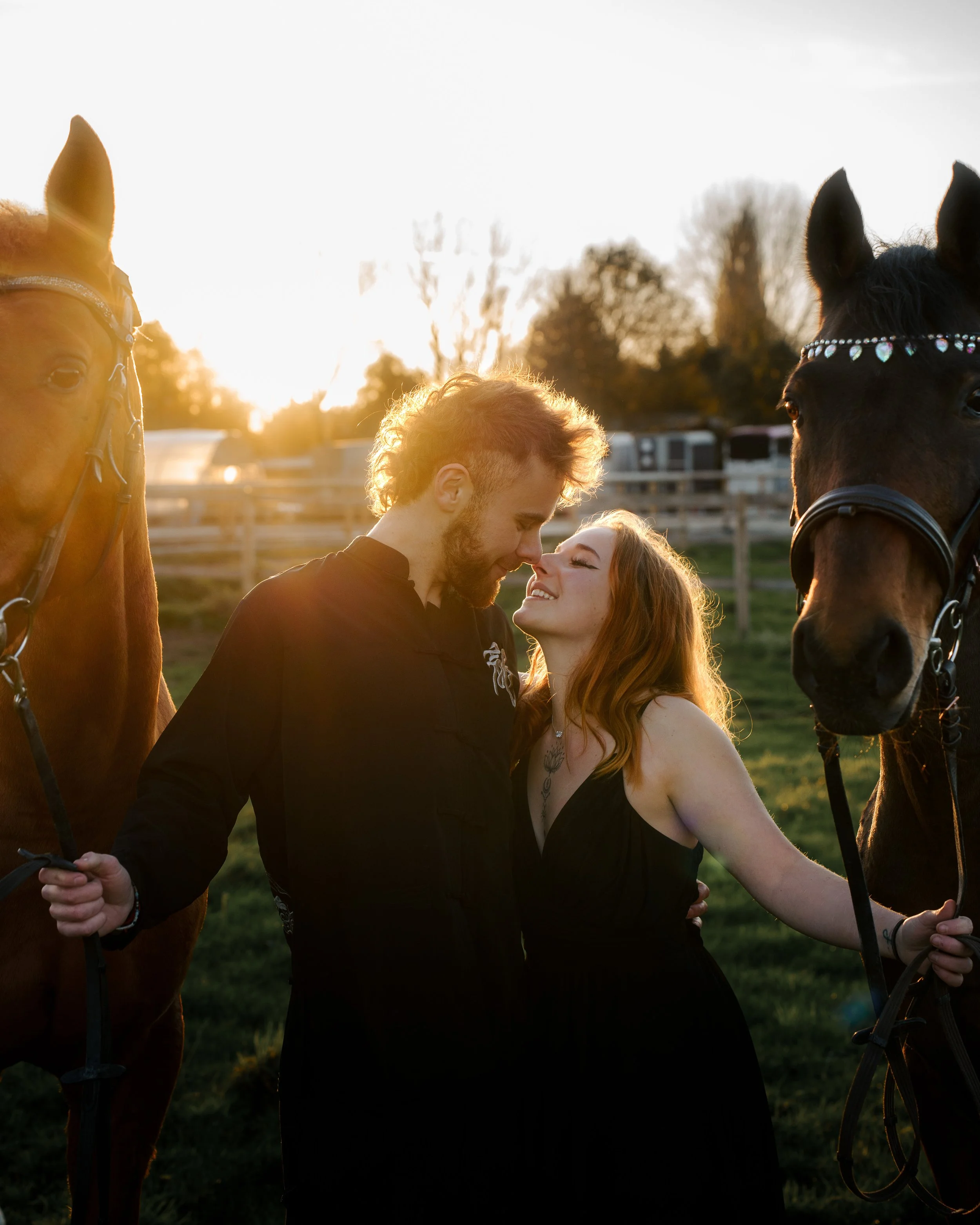 Un couple souriant se tient près de deux chevaux dans un pré lors d'un coucher de soleil.