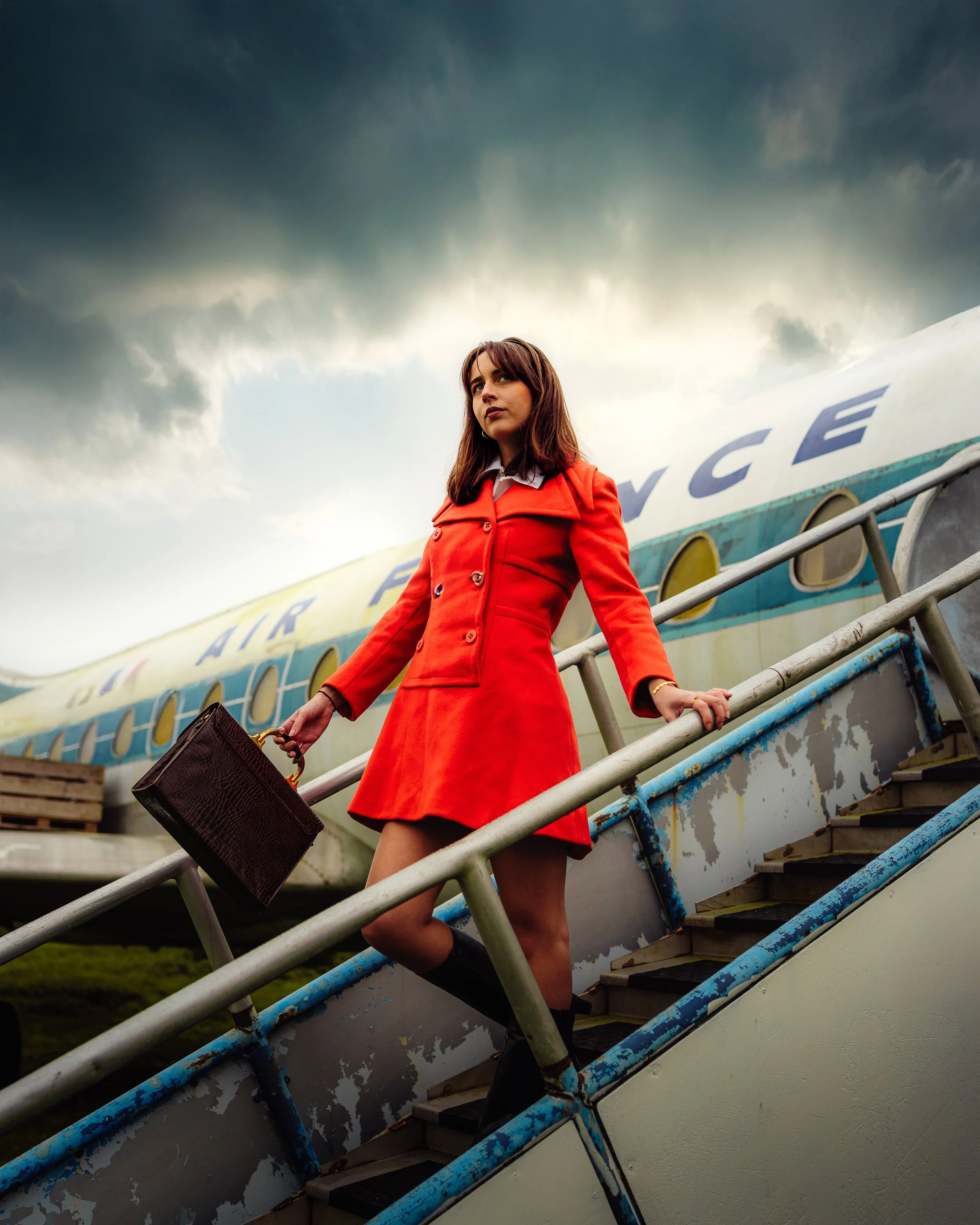 Femme descendant d'un avion en portant un sac à poignée en cuir, portant un manteau rouge, sous un ciel nuageux.