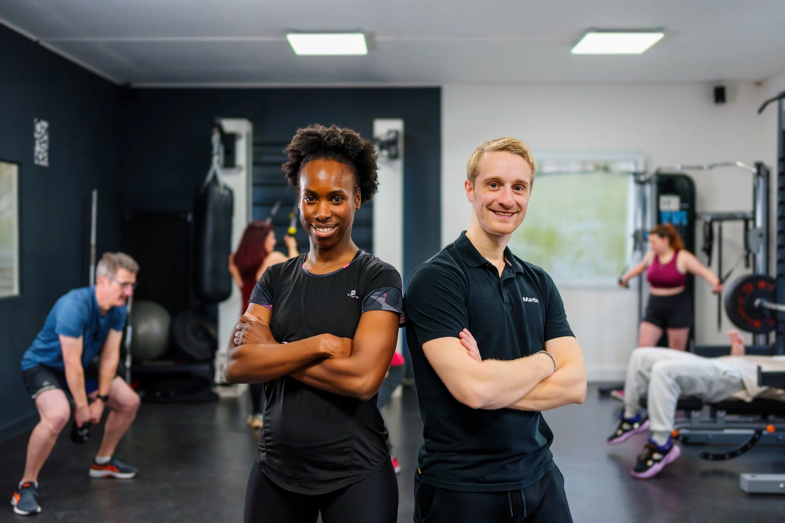 Deux entraîneurs, un homme et une femme, posent avec confiance dans une salle de sport, avec des personnes s'entraînant en arrière-plan.