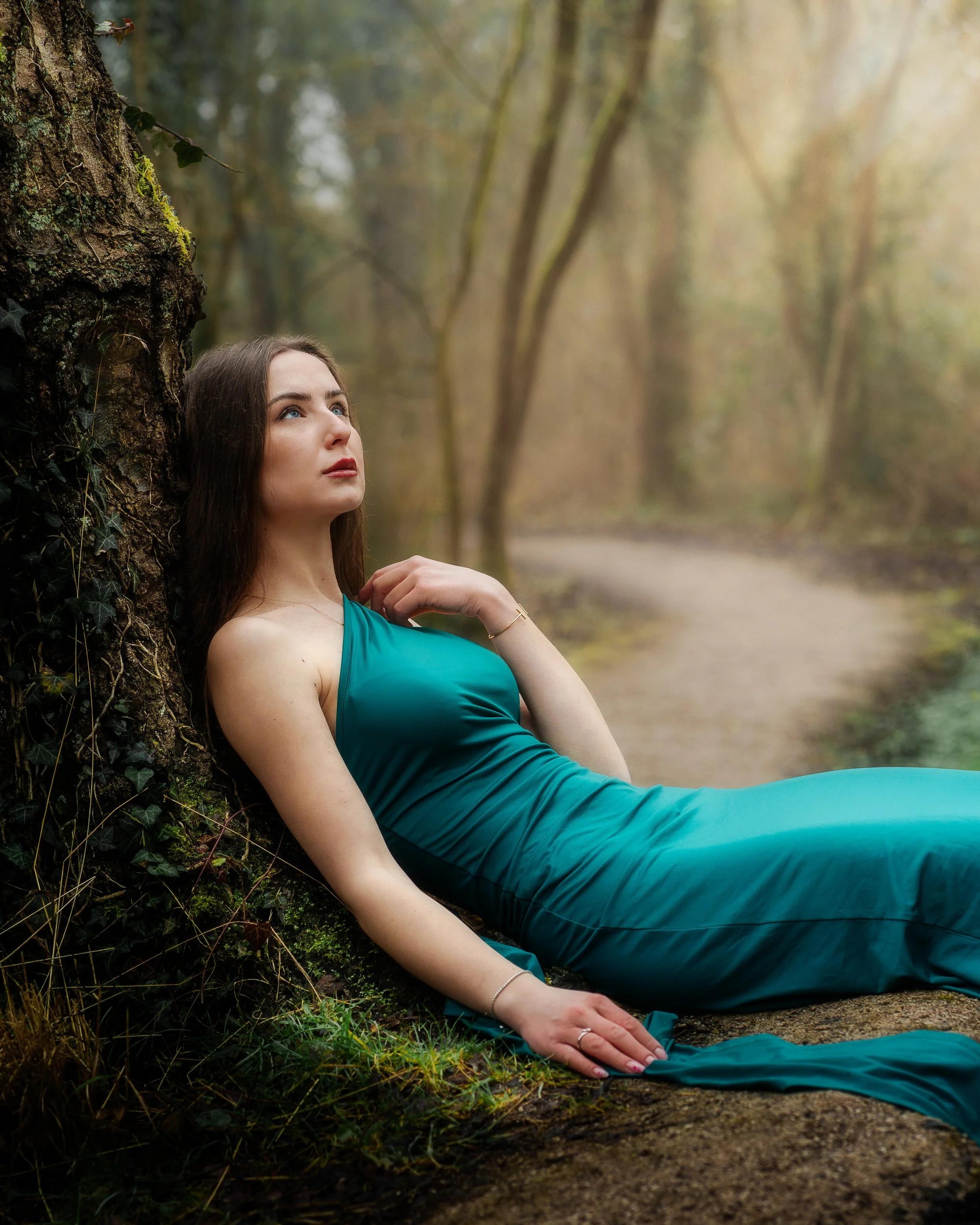 Une femme en robe bleue repose contre un arbre dans une forêt, avec un chemin en arrière-plan.