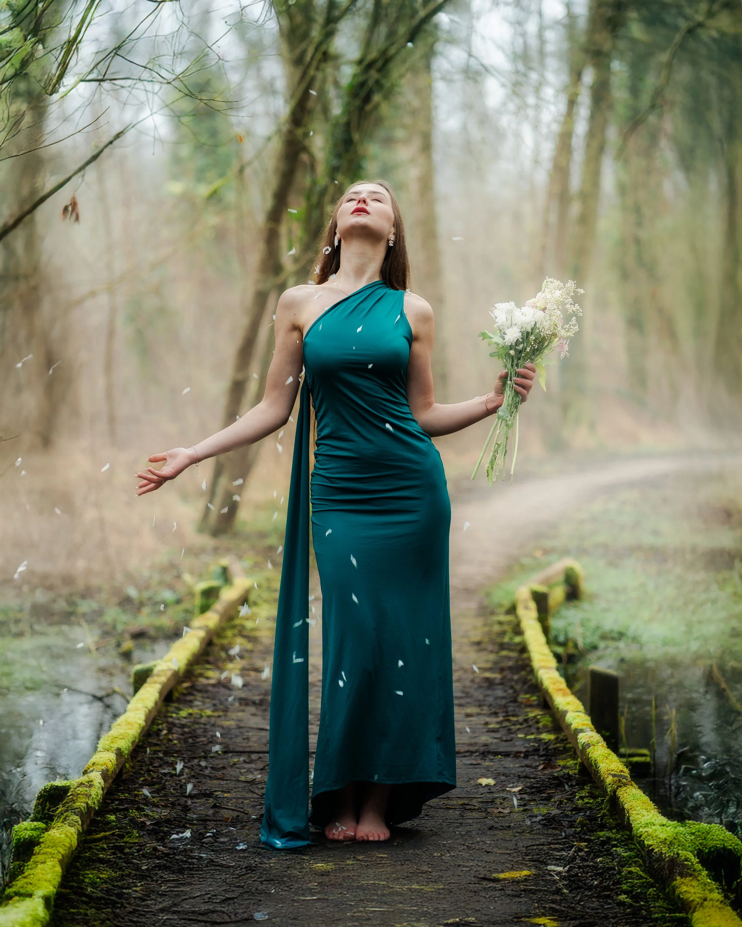 Une femme en robe bleue se tient nue sur un petit pont en bois dans une forêt, tenant un bouquet de fleurs, avec des pétales qui volent dans l'air.