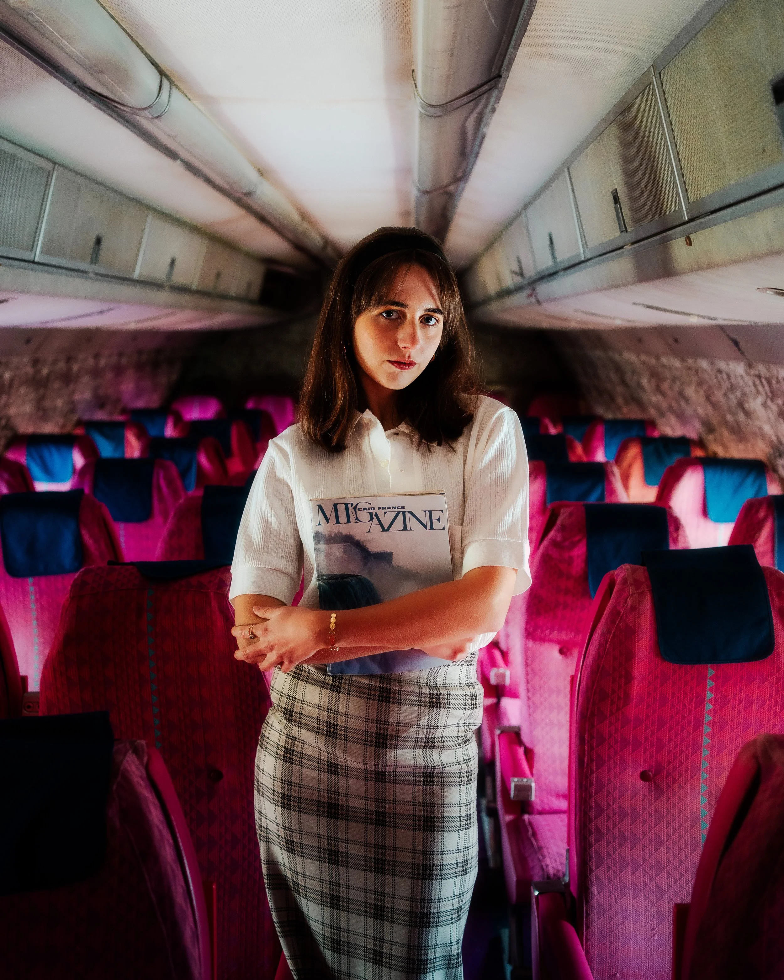 Une femme debout dans un avion, tenant un magazine, avec des sièges rouges et des murs en arrière-plan.