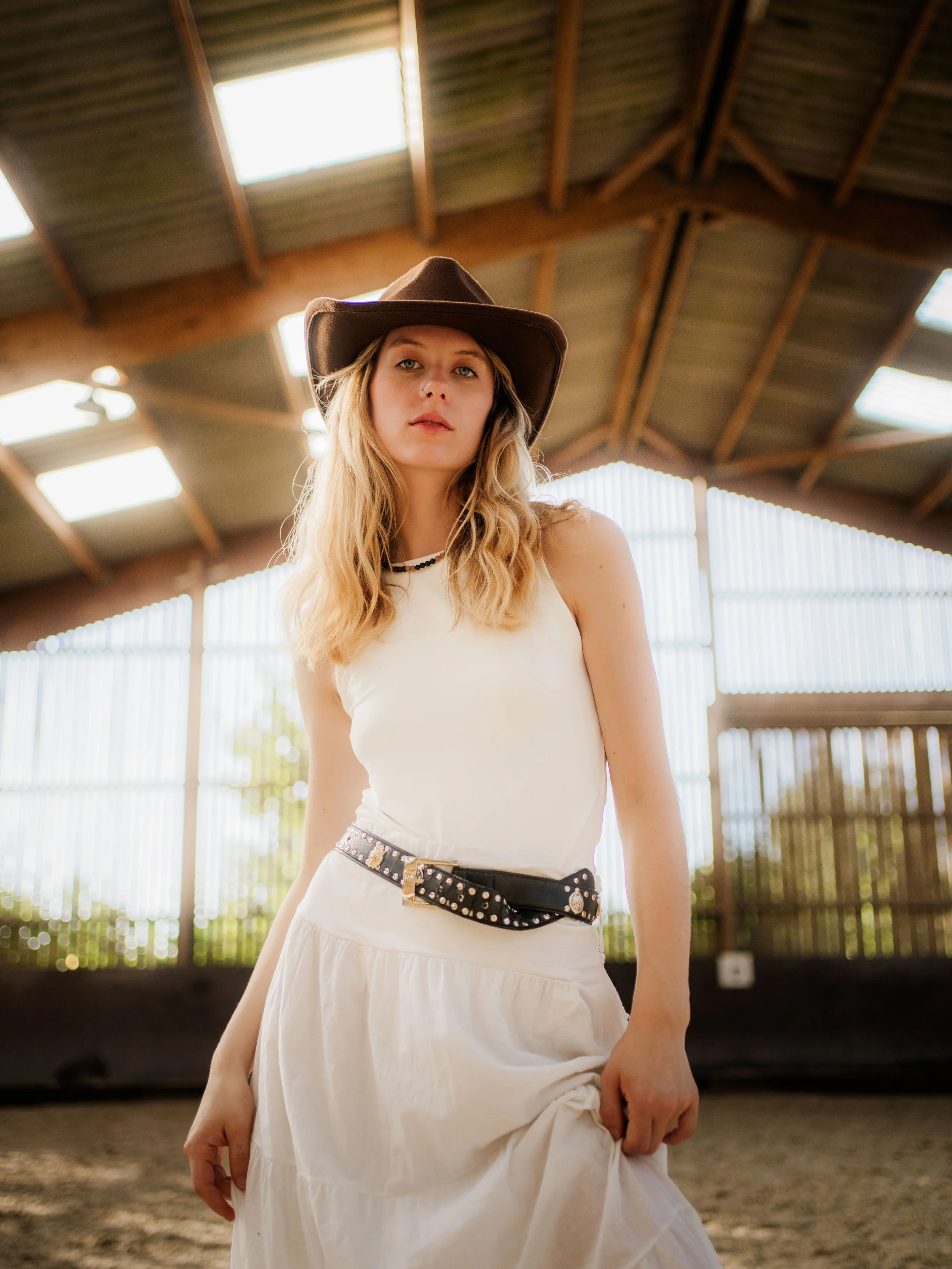Jeune femme portant un chapeau de cowboy, une robe blanche et une ceinture à clous, dans une écurie ou un espace couvert avec un plafond en bois.