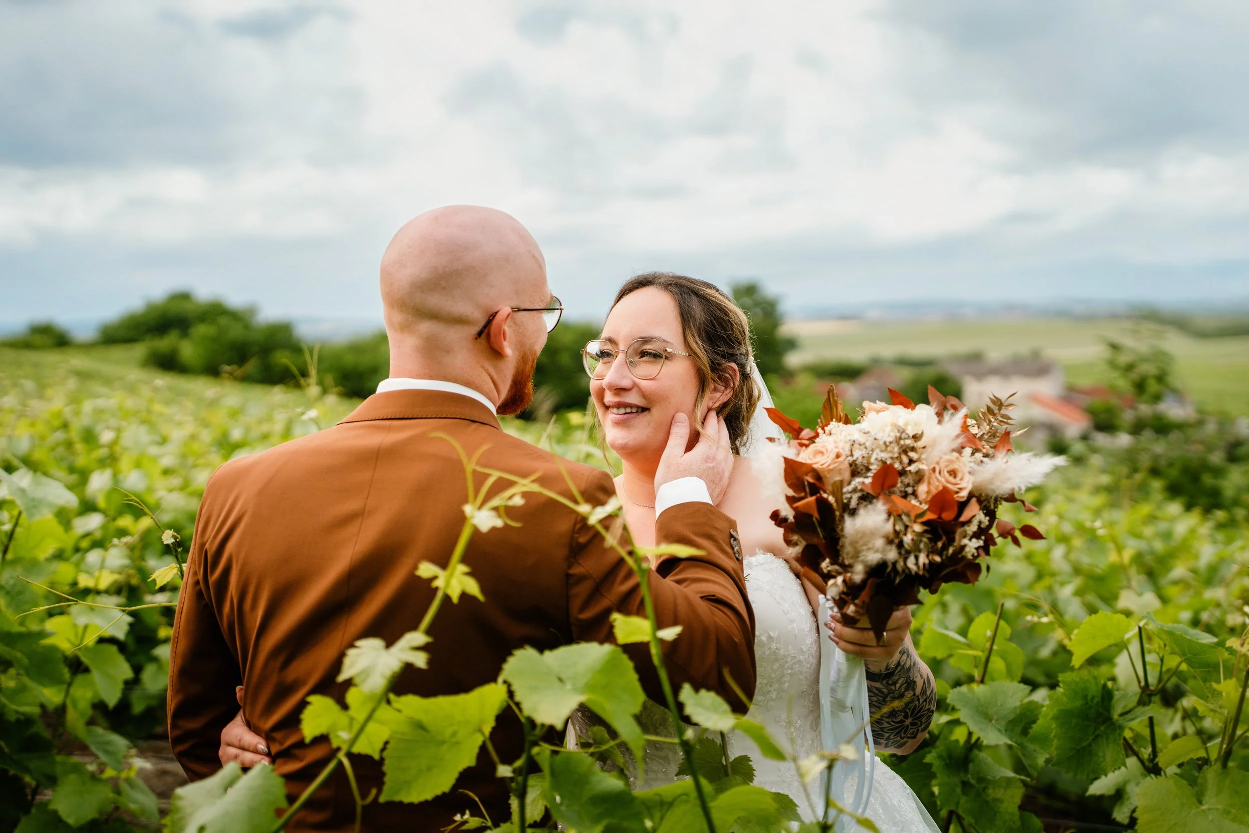 Un couple de mariés dans un champ de vignes, la femme tenue par le marié, souriant et tenant un bouquet de fleurs dans la main.