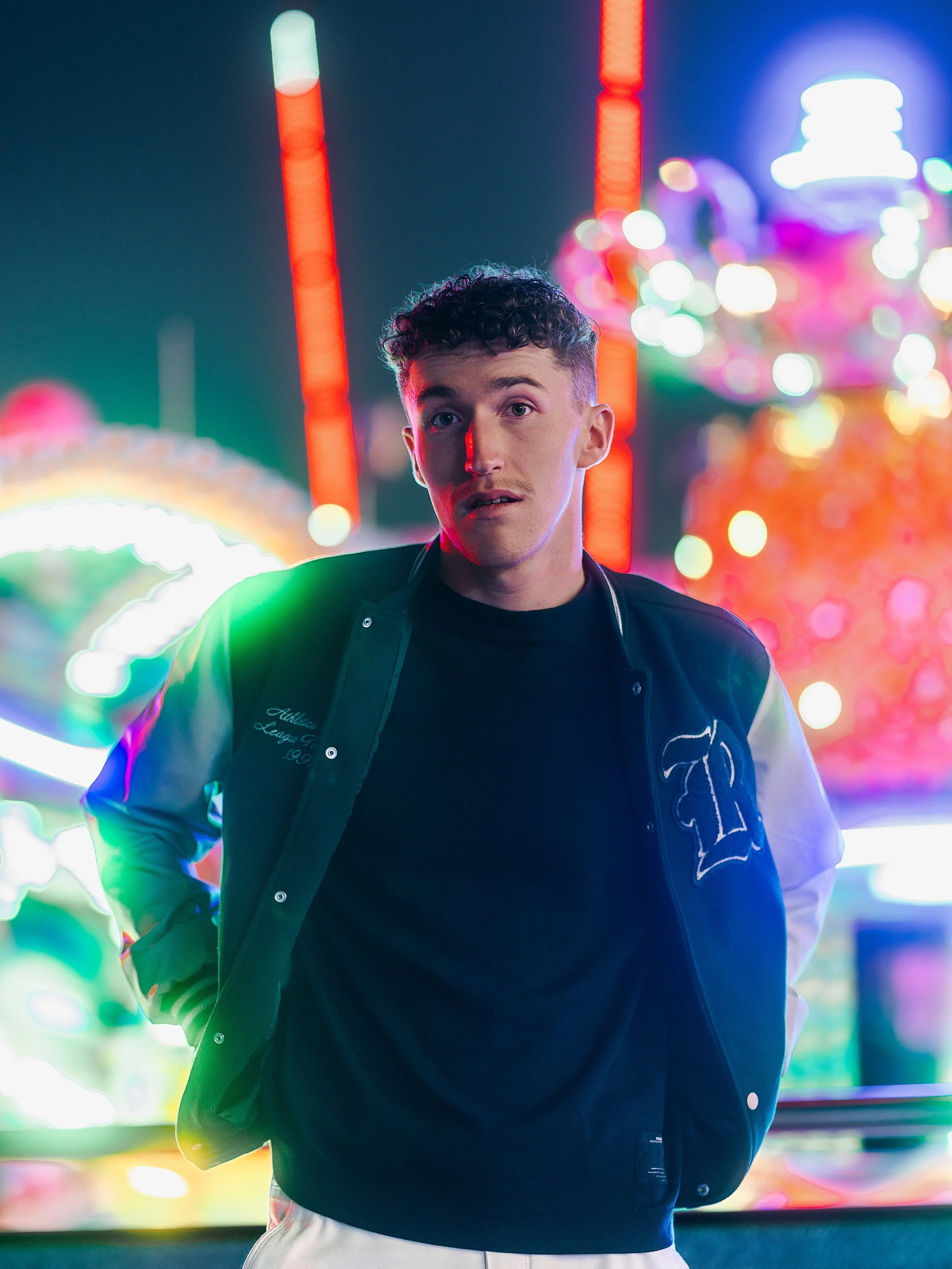 Un jeune homme debout devant une fête foraine illuminée la nuit, avec des lumières colorées de manèges en arrière-plan.