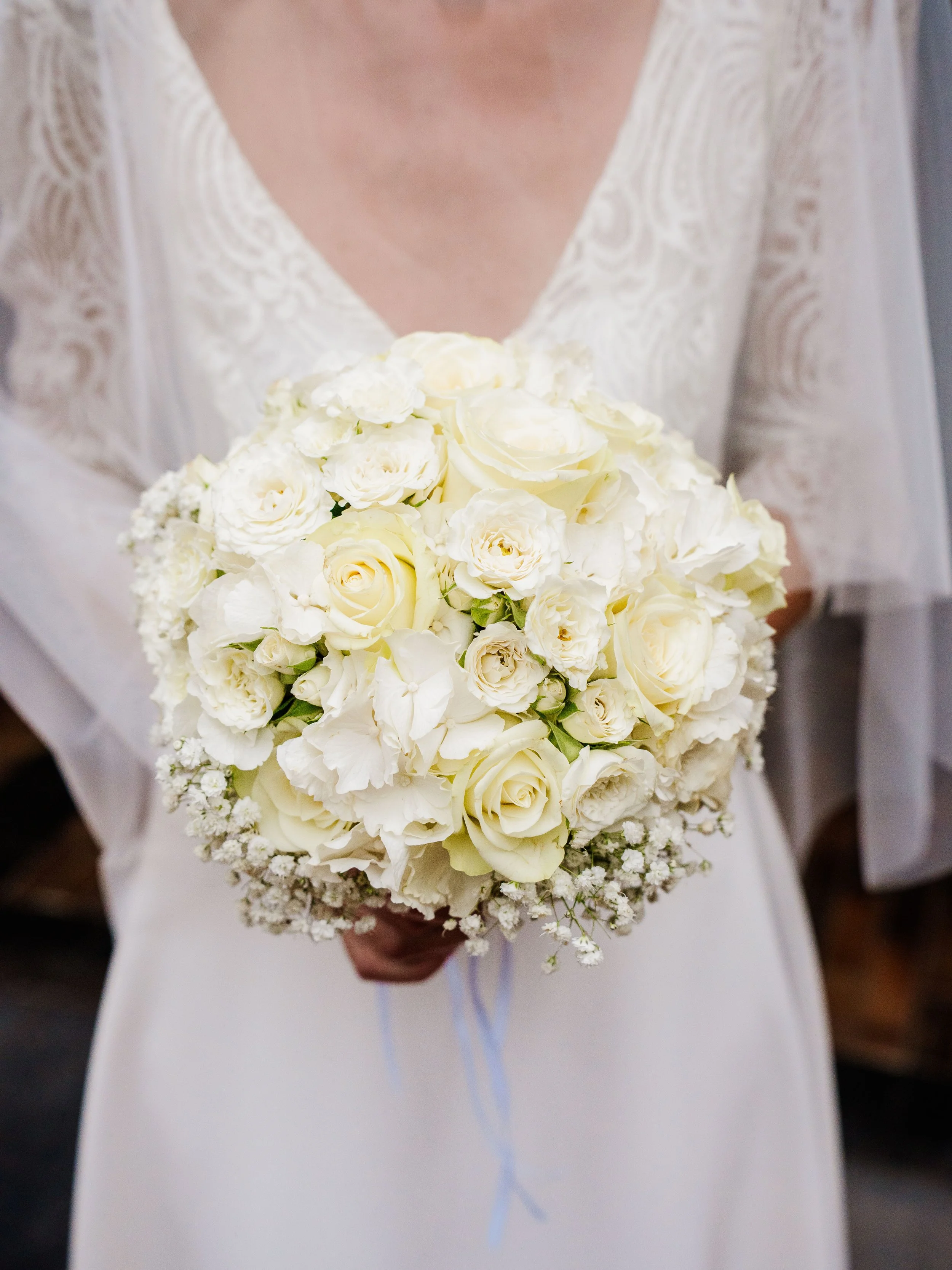Person en robe blanche tenant un bouquet de fleurs blanches, comprenant des roses et des lis, avec un nœud en ruban blanc.