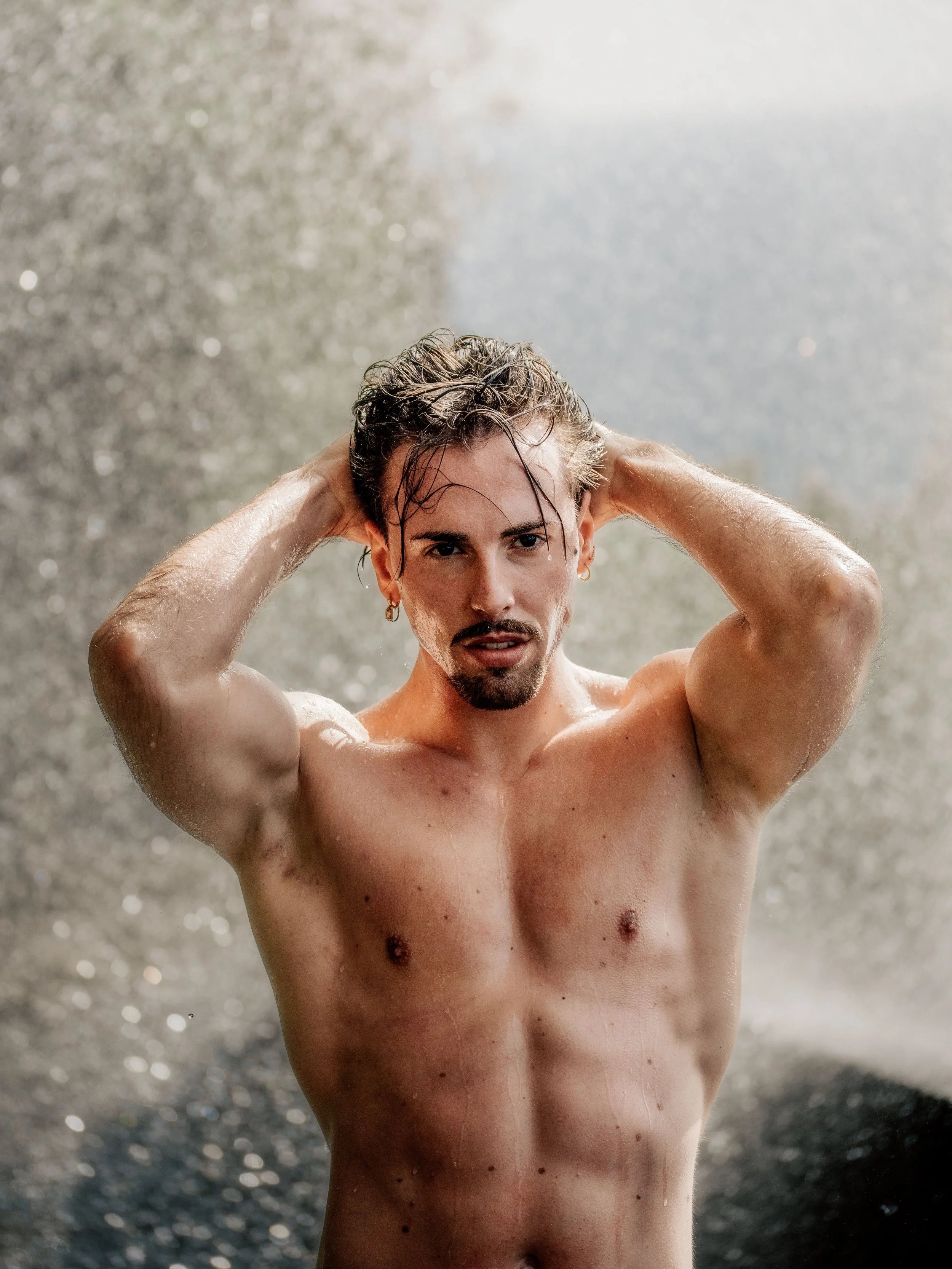 Un homme nu avec les mains dans les cheveux, sous une cascade d'eau, regardant vers l'avant.