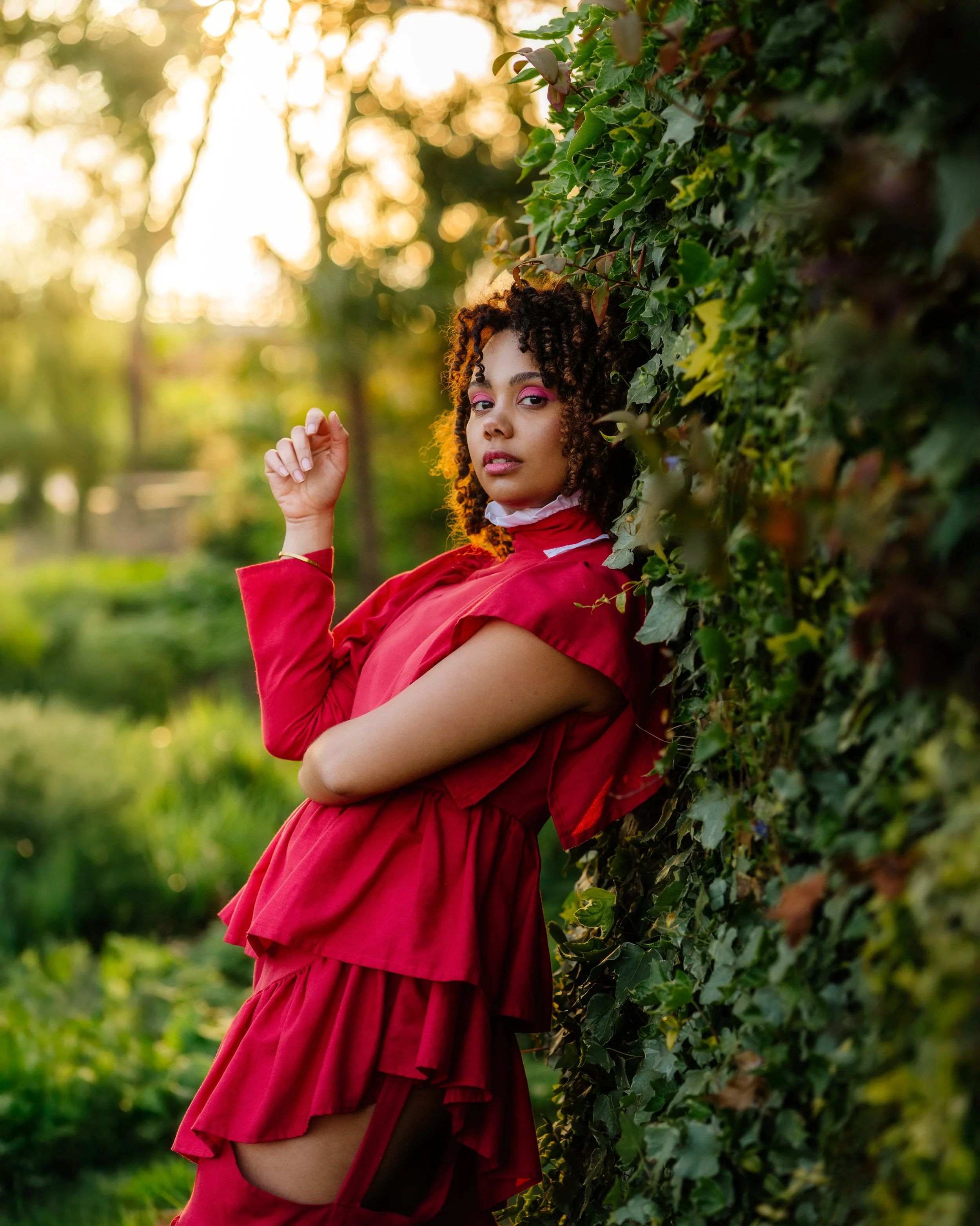 Jeune femme africaine portant une robe rouge, posant contre un mur recouvert de lierre dans un parc au coucher du soleil, regardant l'objectif.