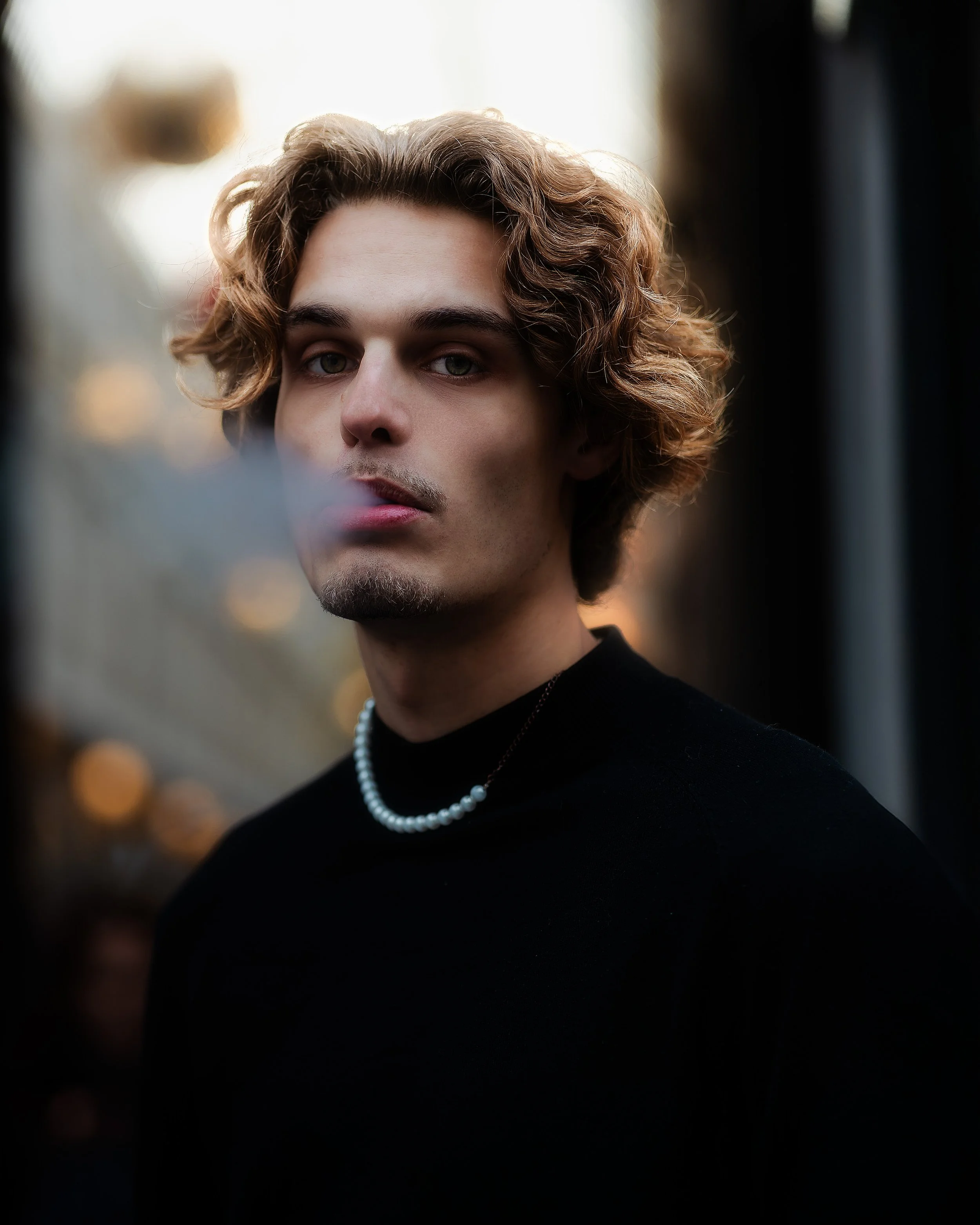 Jeune homme avec cheveux bouclés, portant un t-shirt noir et un collier de perles, regardant vers l'objectif, avec un flou artistique en arrière-plan