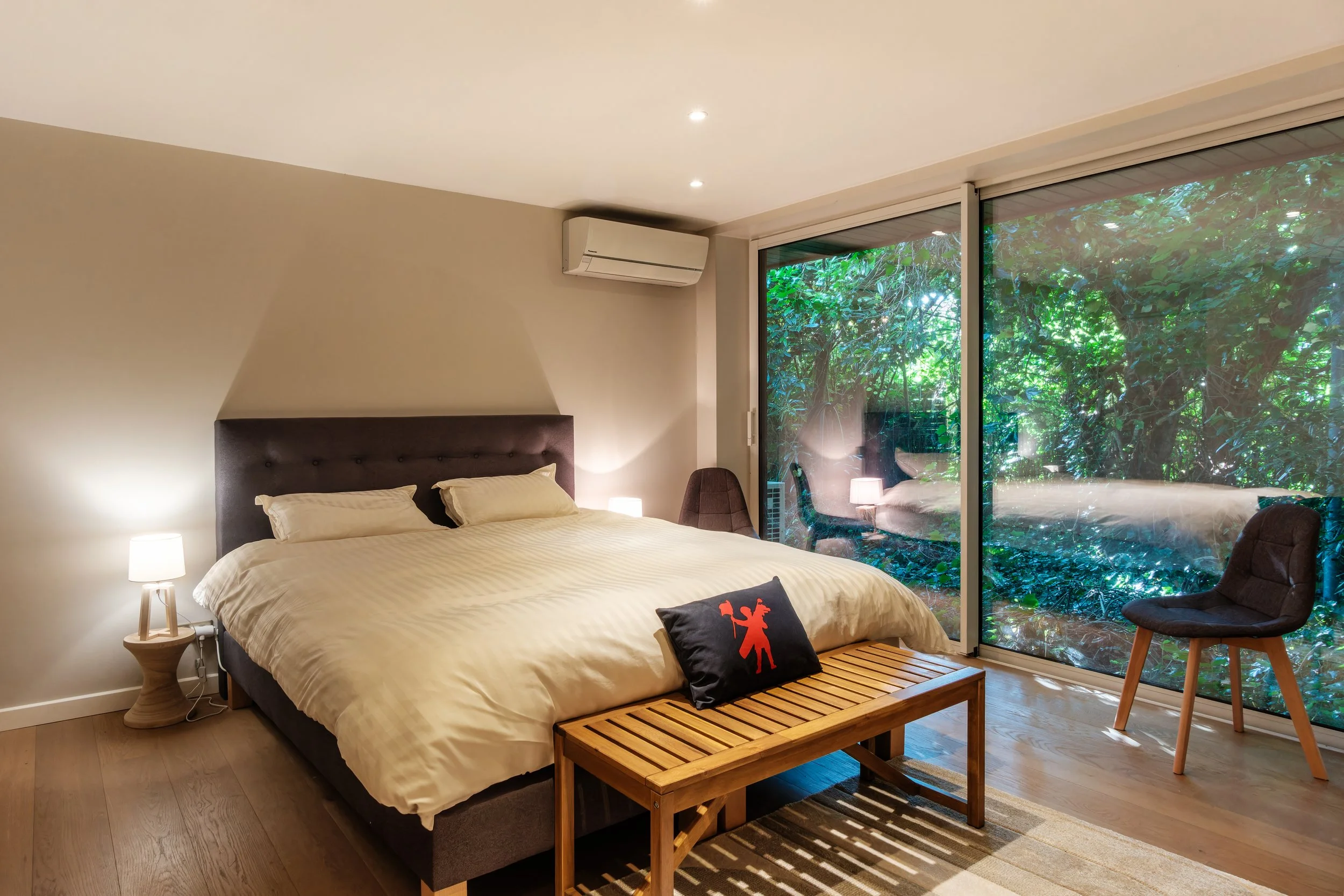 Chambre moderne avec grand lit à draps beiges, coussins, lampe de chevet, fauteuil et banc en bois, grande fenêtre donnant sur un espace vert