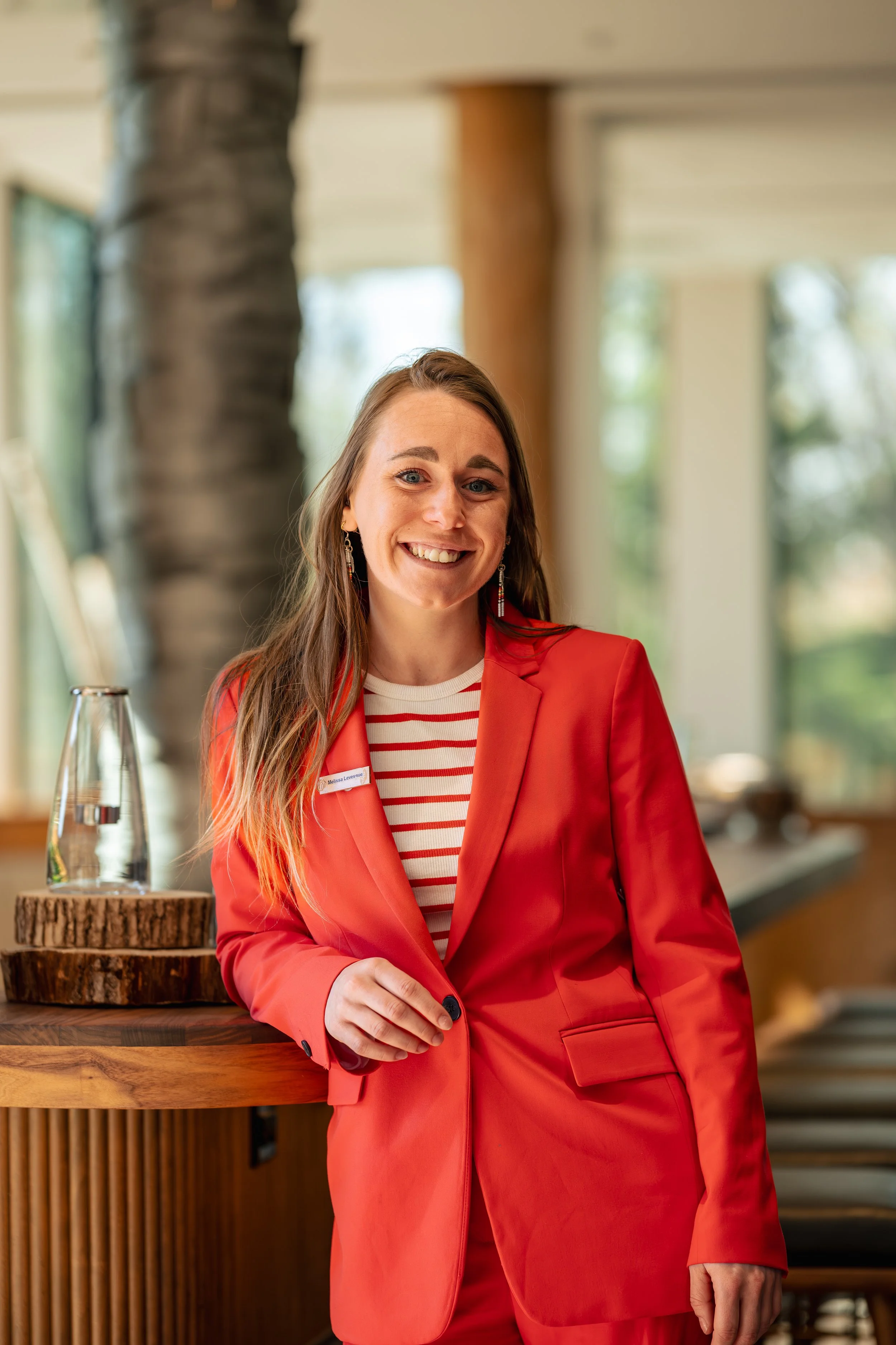 Une femme souriante en costume rouge, portant une chemise à rayures, se tient près d'un comptoir en bois avec un verre d'eau. L'environnement semble être intérieur, avec de grandes fenêtres et un décor en bois.