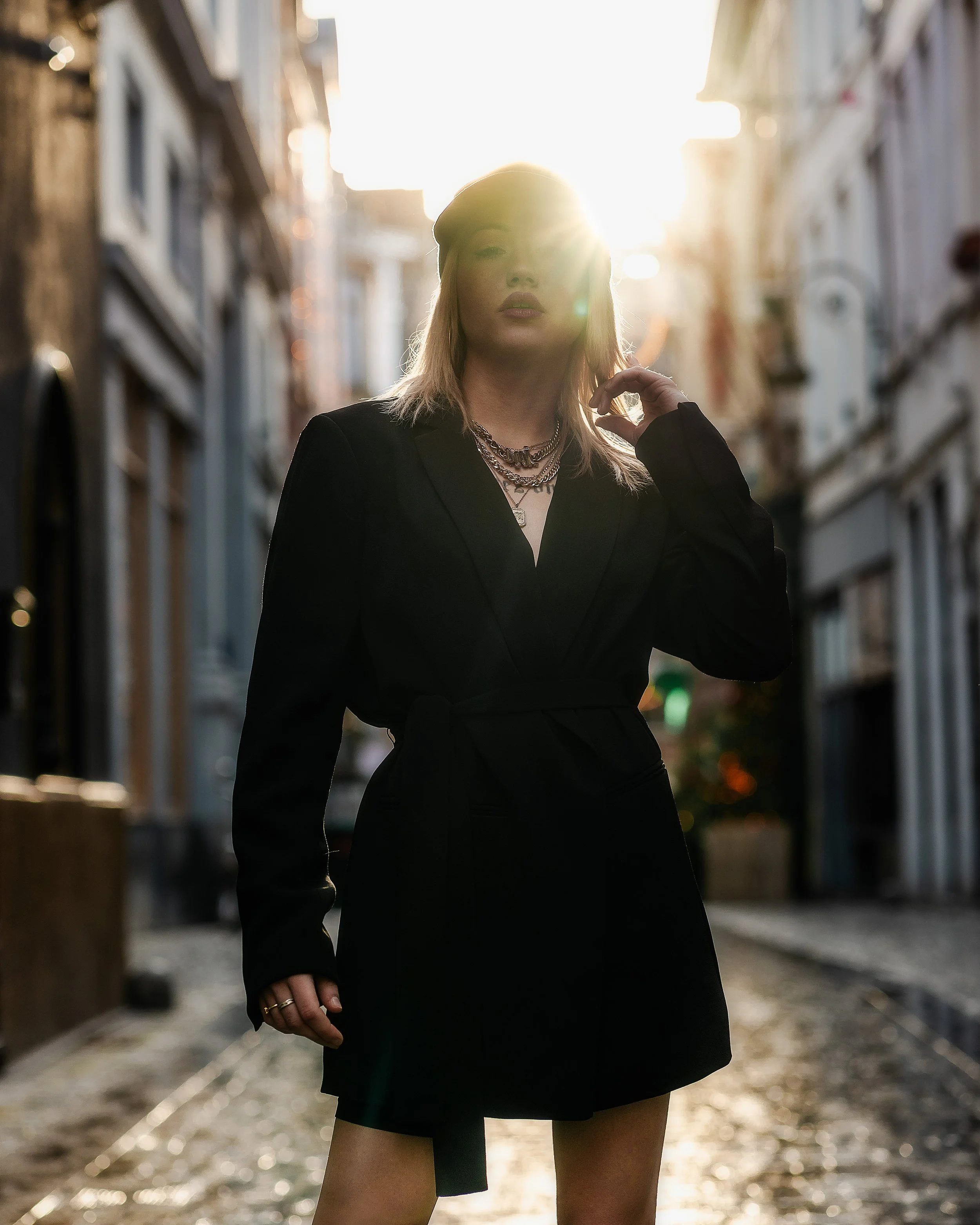 Jeune femme posant dans une rue ensoleillée, vêtue d'un blazer noir, portant des colliers et une bague, avec le soleil derrière elle.