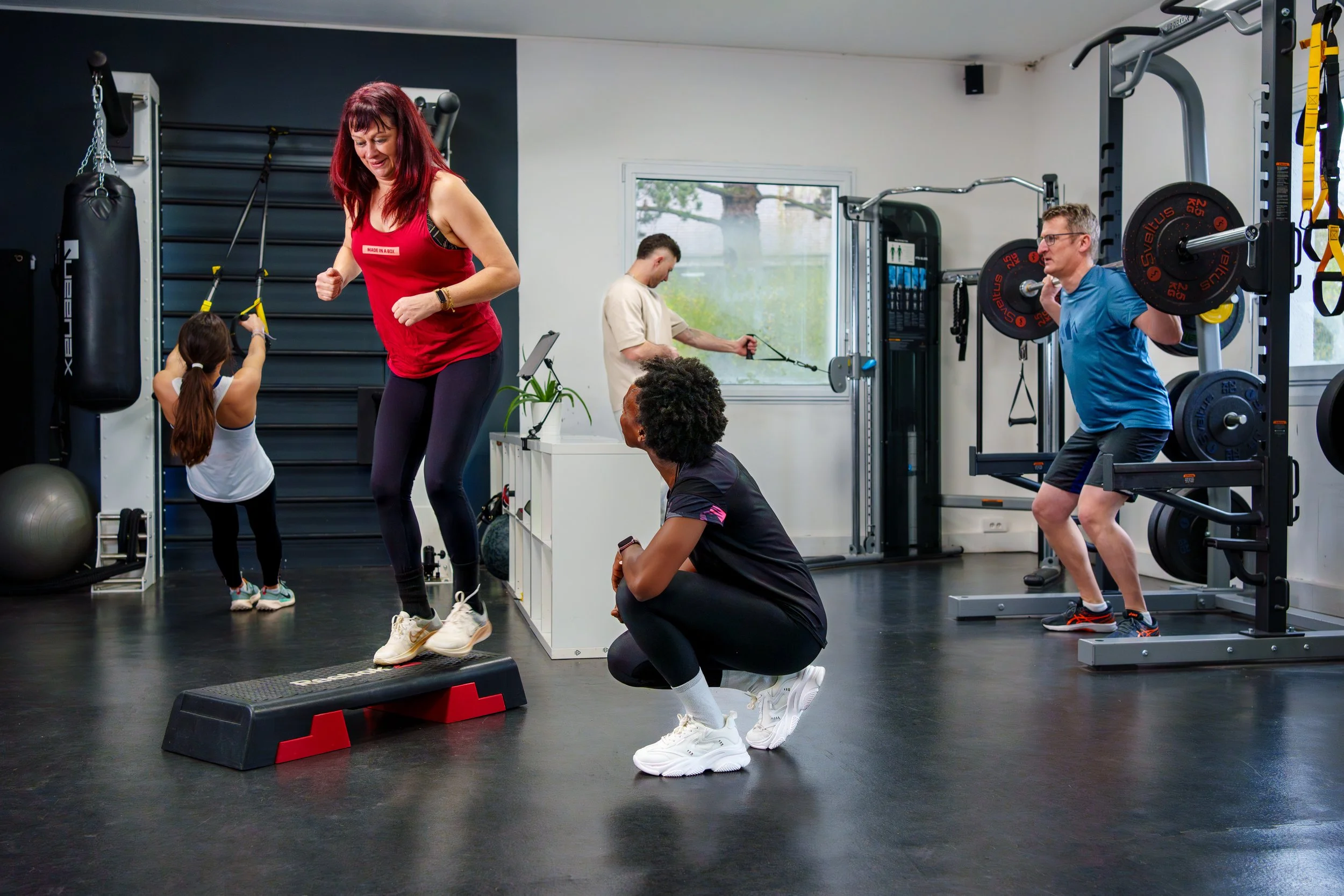Groupe de personnes s'entraînant dans une salle de sport, avec un homme soulevant des poids, une femme sautant à la corde, une femme s'entraînant avec un step, et deux autres personnes utilisant diverses machines.