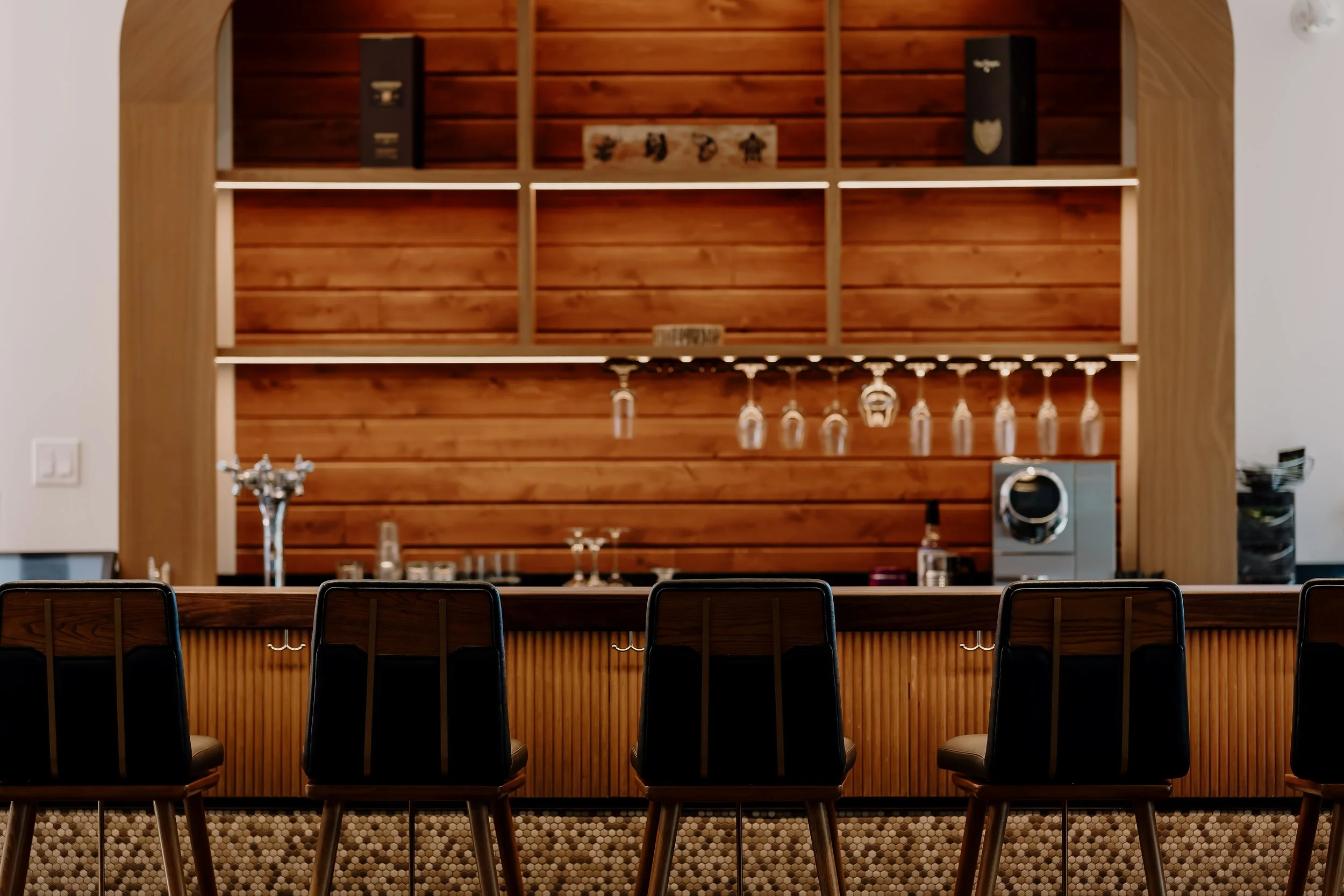 Un bar avec un comptoir en bois et des chaises en acier noir, décor en bois chaud et étagères avec verres suspendus.