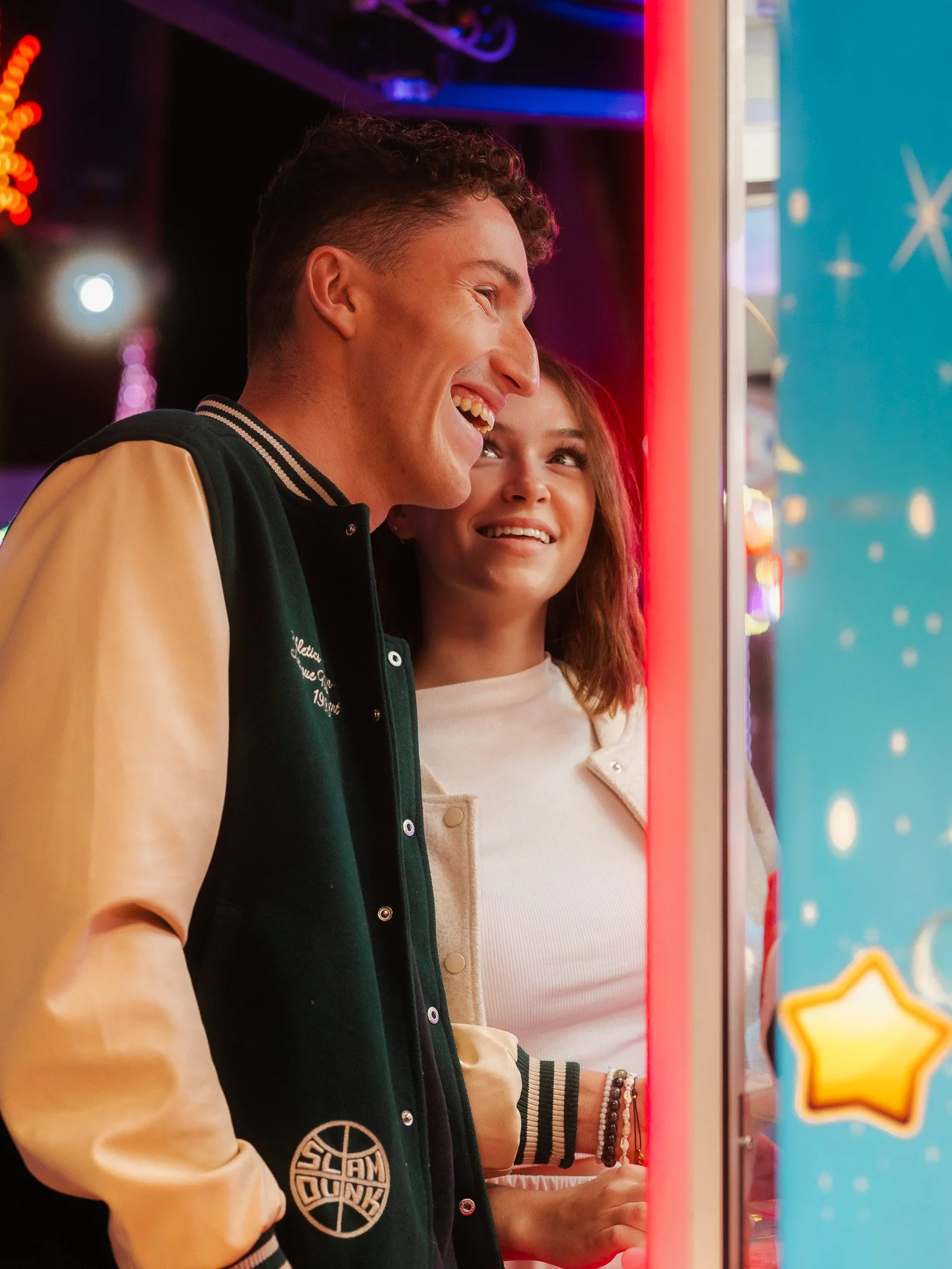 Un homme et une femme sourient en regardant une machine de jeux d'arcade lumineuse dans une salle de jeux ou un centre de divertissement, avec des lumières colorées en arrière-plan.