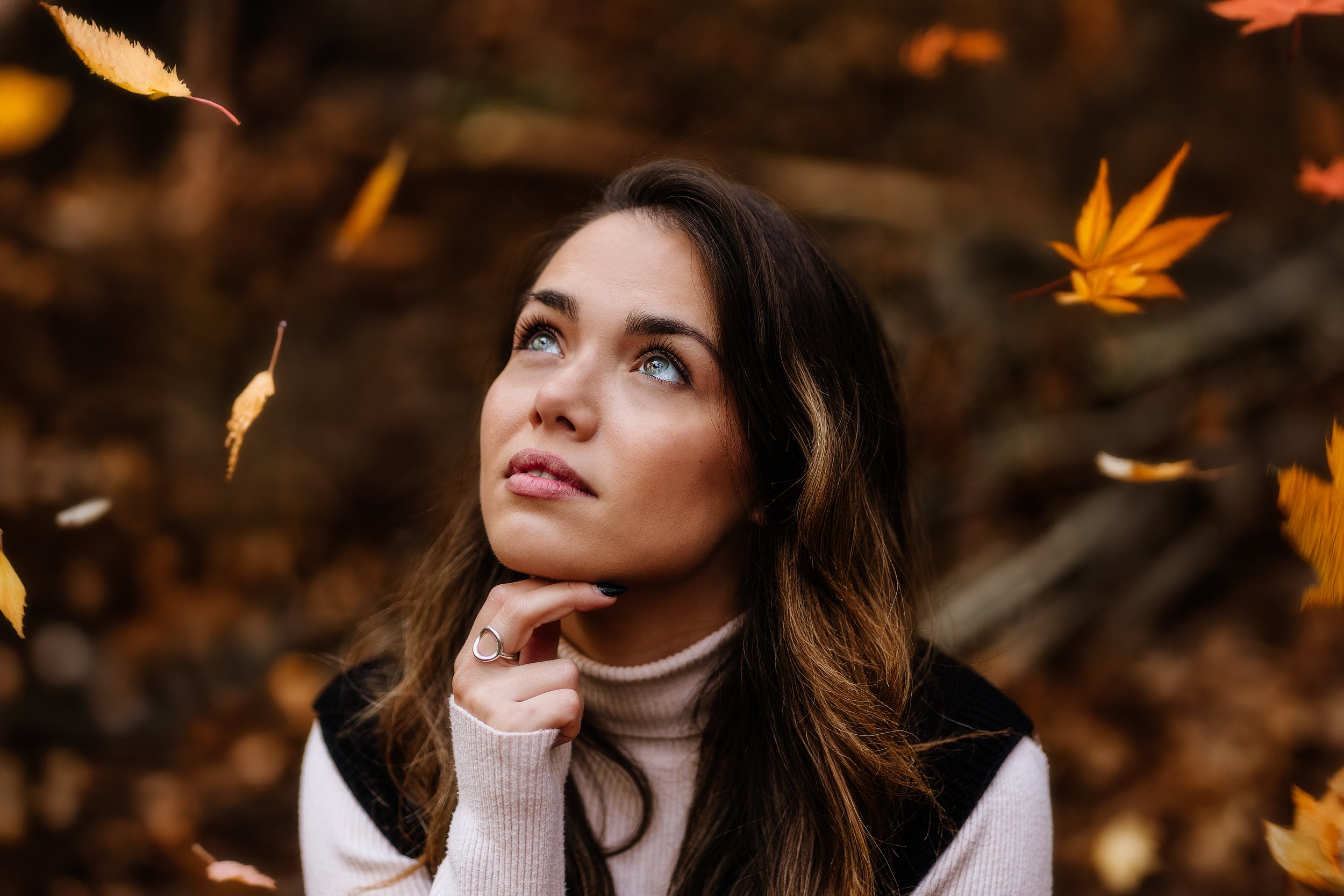 Une jeune femme avec des cheveux bruns et des yeux bleus, portant un pull beige à col roulé, penseuse, entourée de feuilles d'automne tombées.