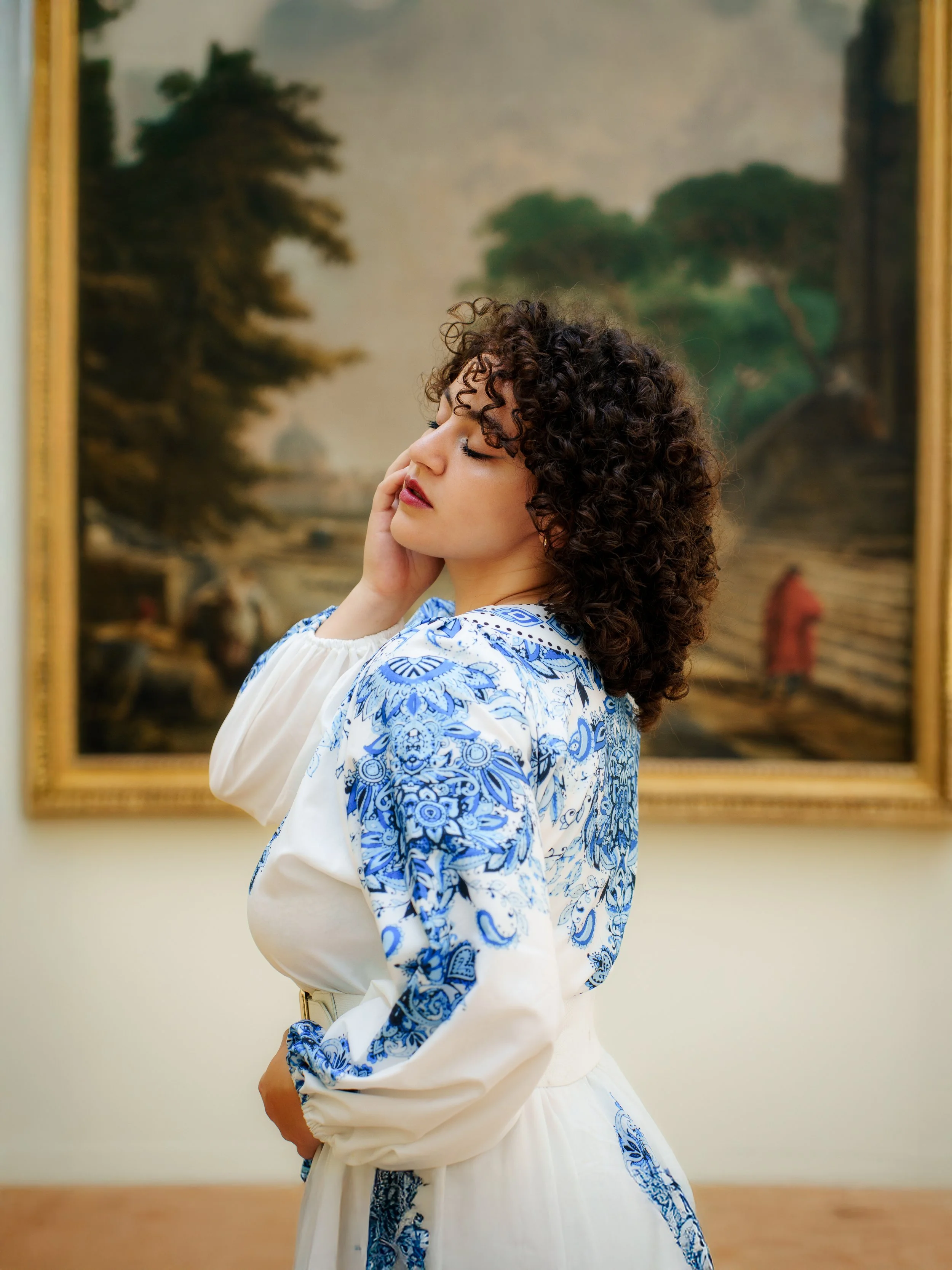Jeune femme aux cheveux bouclés posant dans un musée, vêtue d'une robe blanche avec des motifs bleus, devant un tableau paysager.