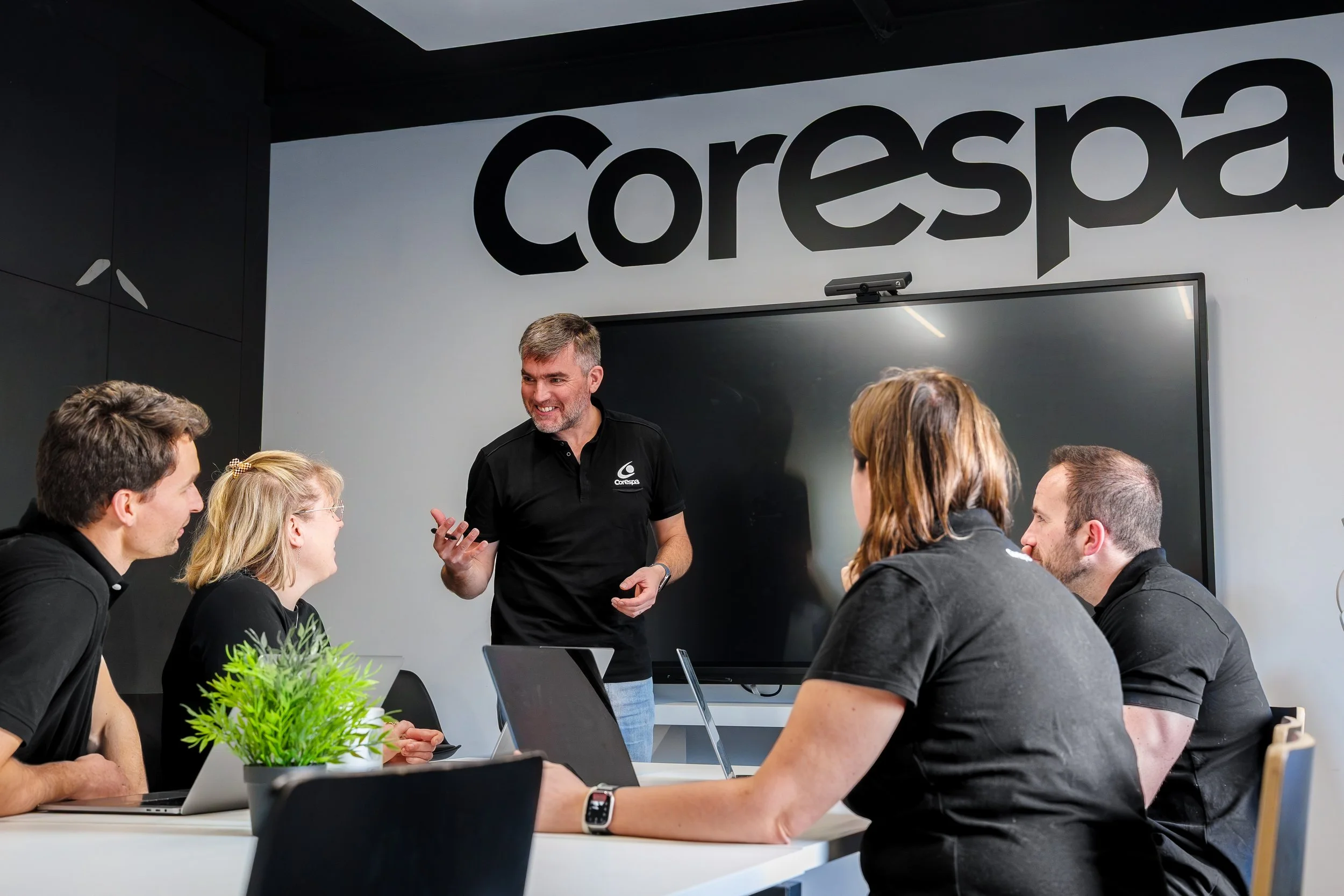 Une personne en train de faire une présentation lors d'une réunion dans une salle de conférence contemporaine, avec un grand écran noir derrière elle et le logo 'Corespa' en arrière-plan.