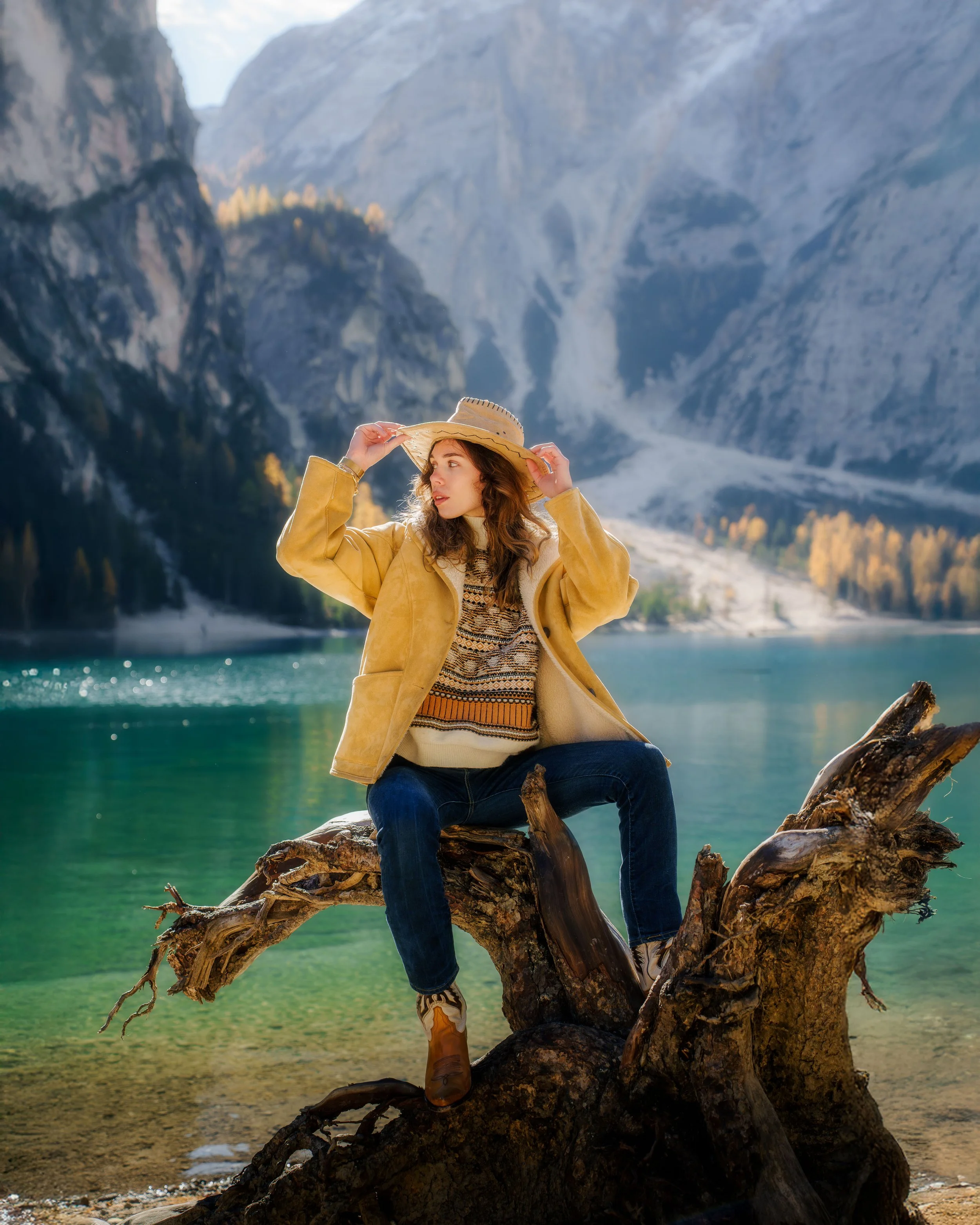 Jeune femme assise sur une branche d'arbre au bord d'un lac montagneux, portant un chapeau, un pull, un manteau jaune et des bottes, avec des montagnes en arrière-plan.