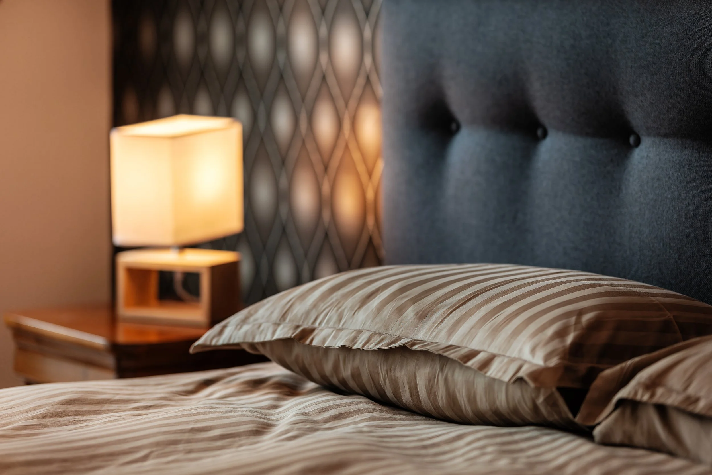 Un lit fait avec une taie d'oreiller et une couverture à rayures beiges, un chevet en bois avec une lampe allumée, et un fond avec un mur noir à motifs en forme de diamants.