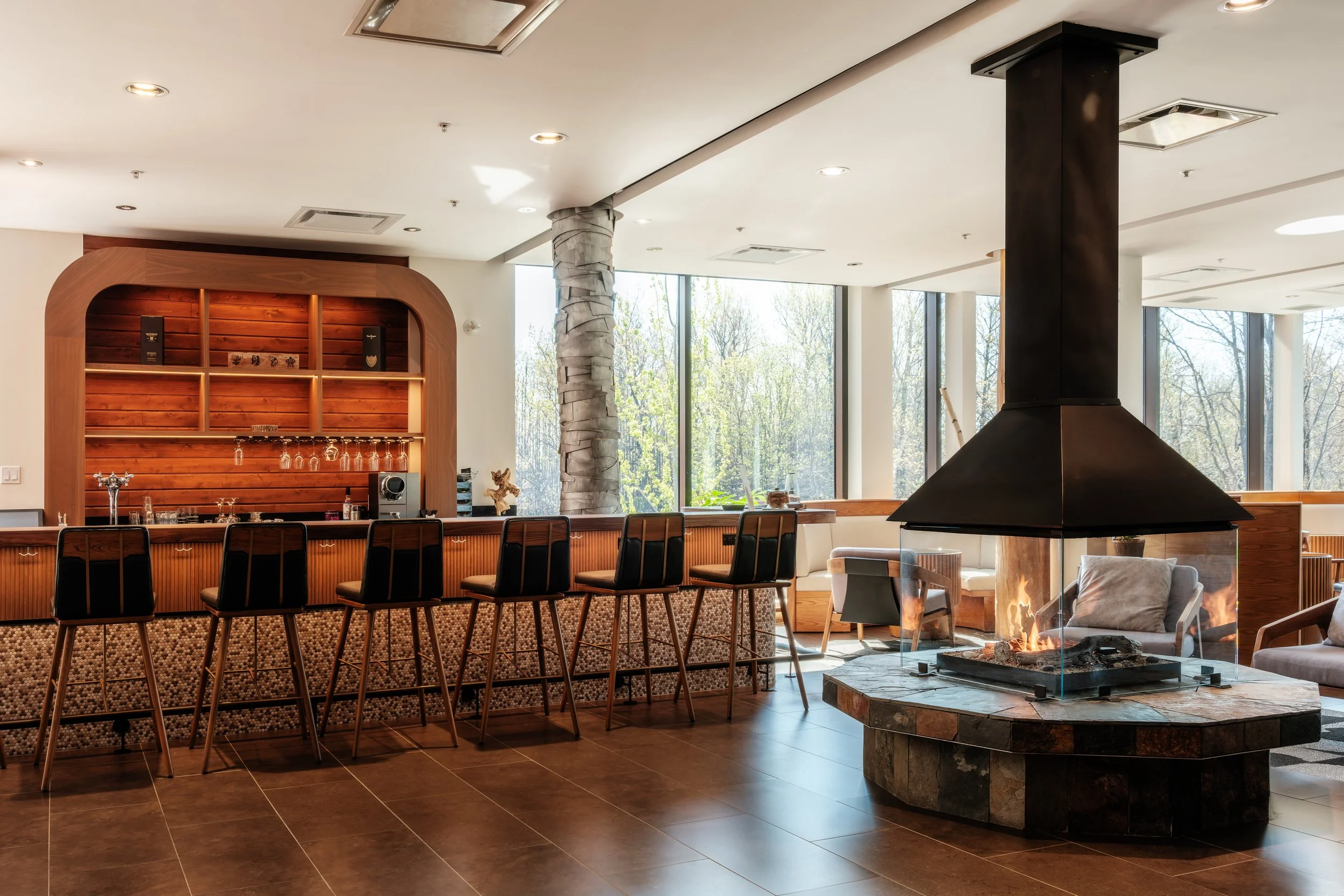 Intérieur d'un bar dans un hôtel ou un restaurant avec un coin cheminée, grandes fenêtres laissant entrer la lumière naturelle, comptoir de bar avec huit chaises et étagère en bois derrière, et un coin salon avec des fauteuils et des tables.