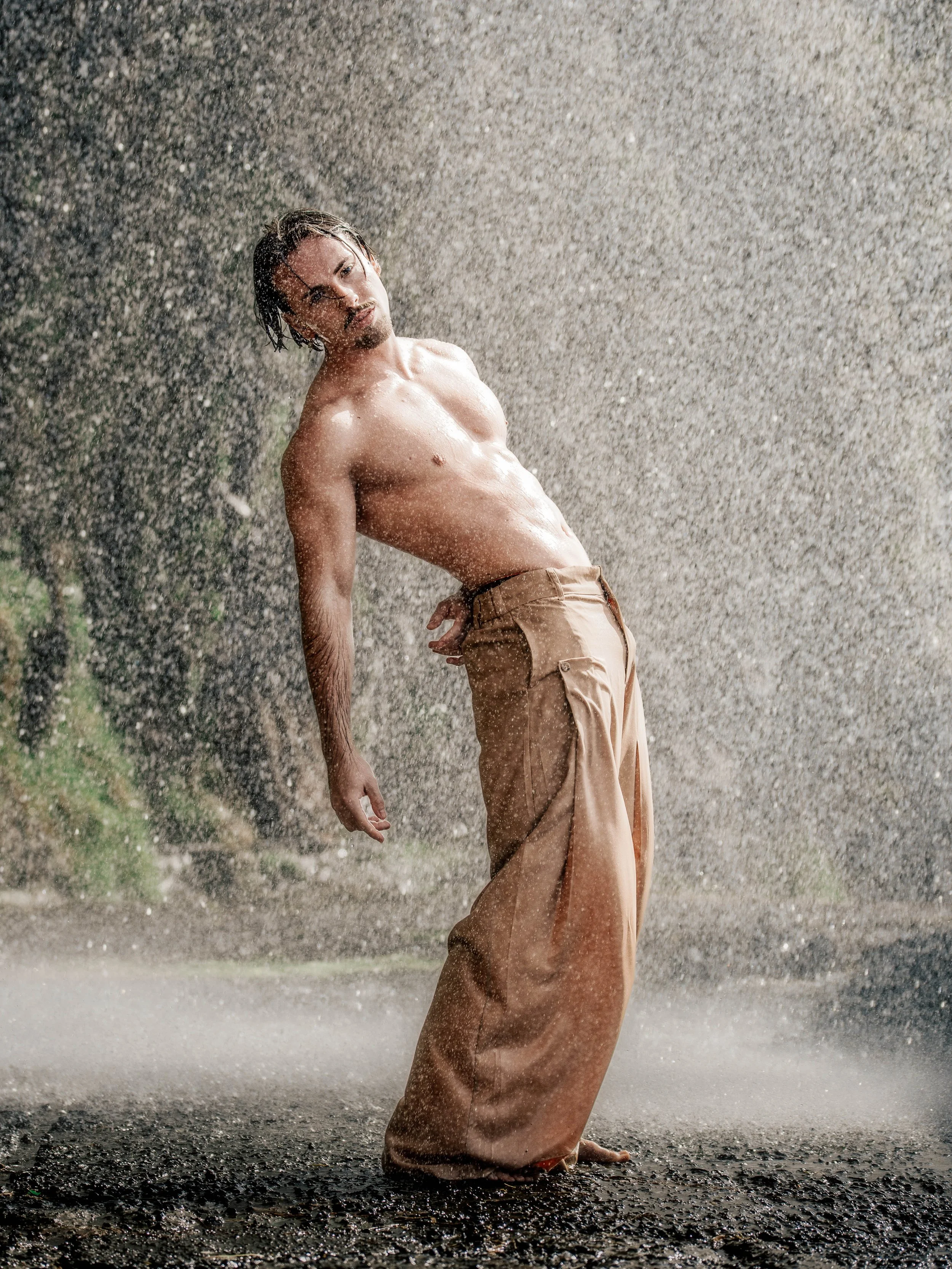 Homme nu debout sous la pluie, portant un pantalon beige, avec une expression méditative dans un environnement naturel.