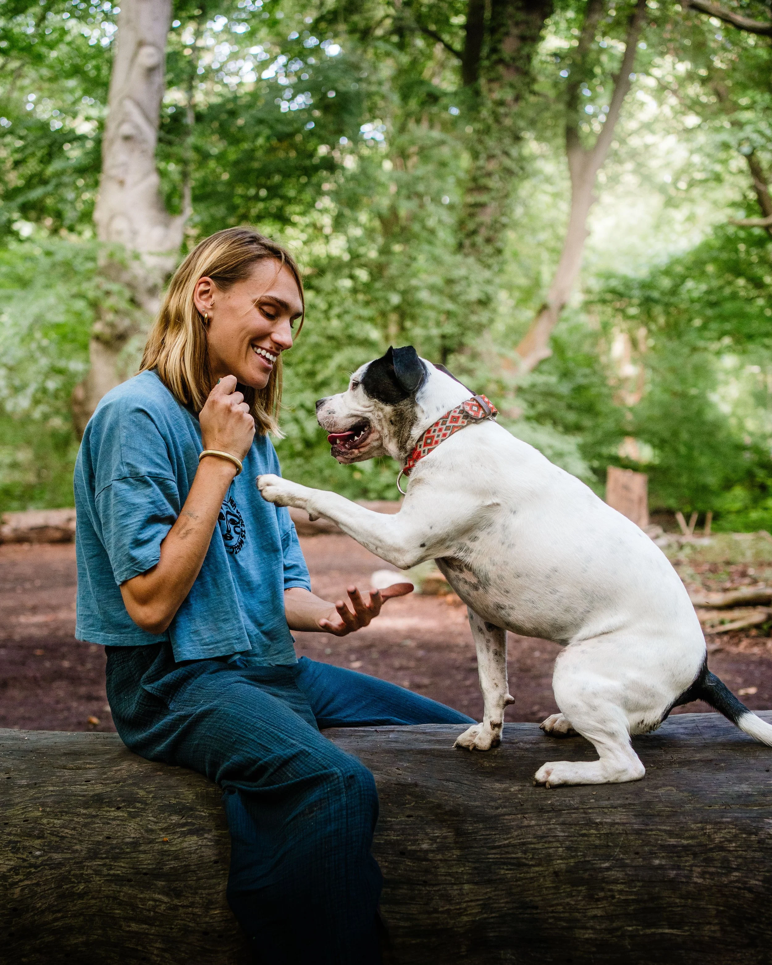 Une femme souriante avec un chien blanc à tache noire en train de jouer dans une forêt verdoyante.