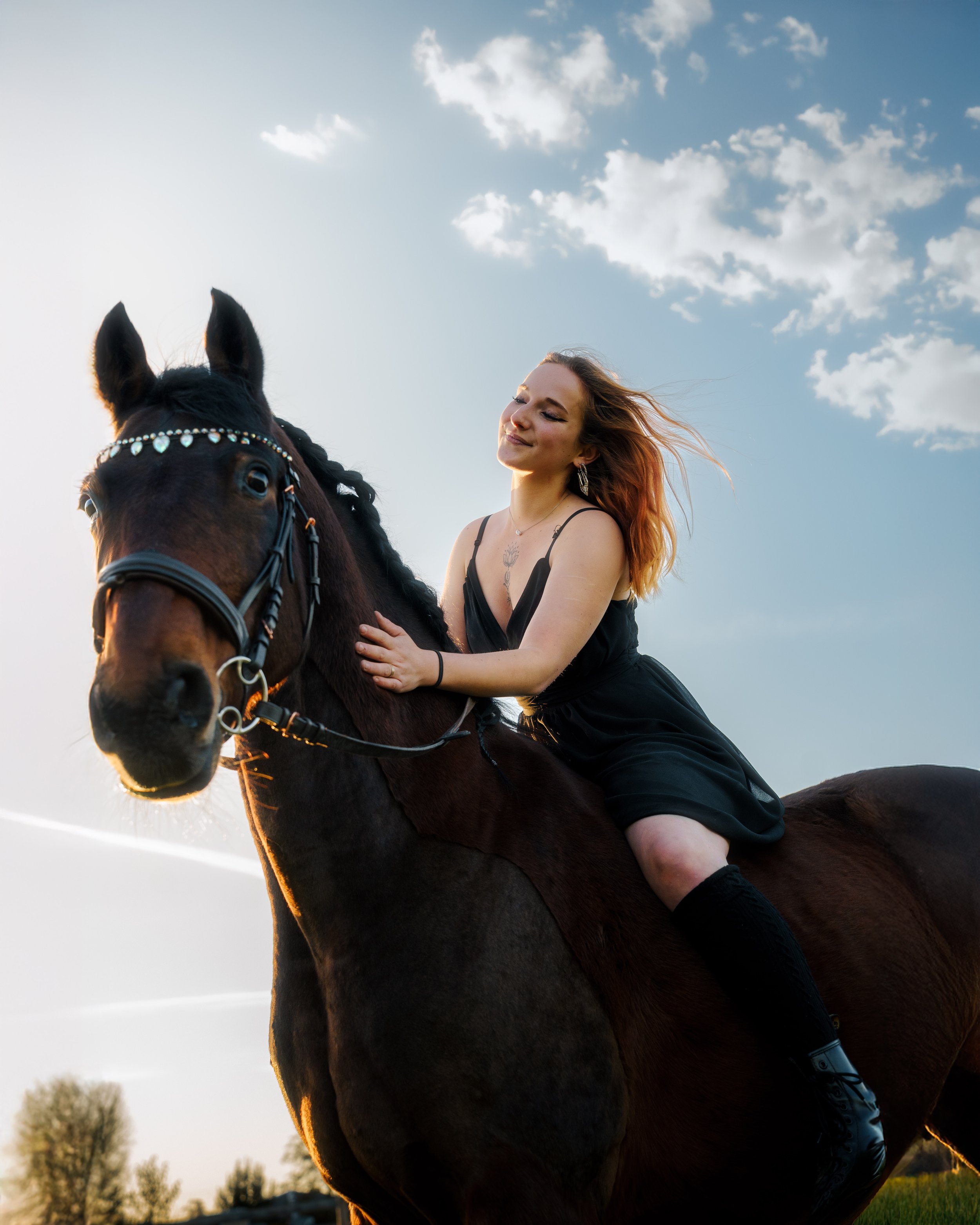 Une femme avec des cheveux rouges assise sur un cheval noir sous un ciel bleu avec quelques nuages.