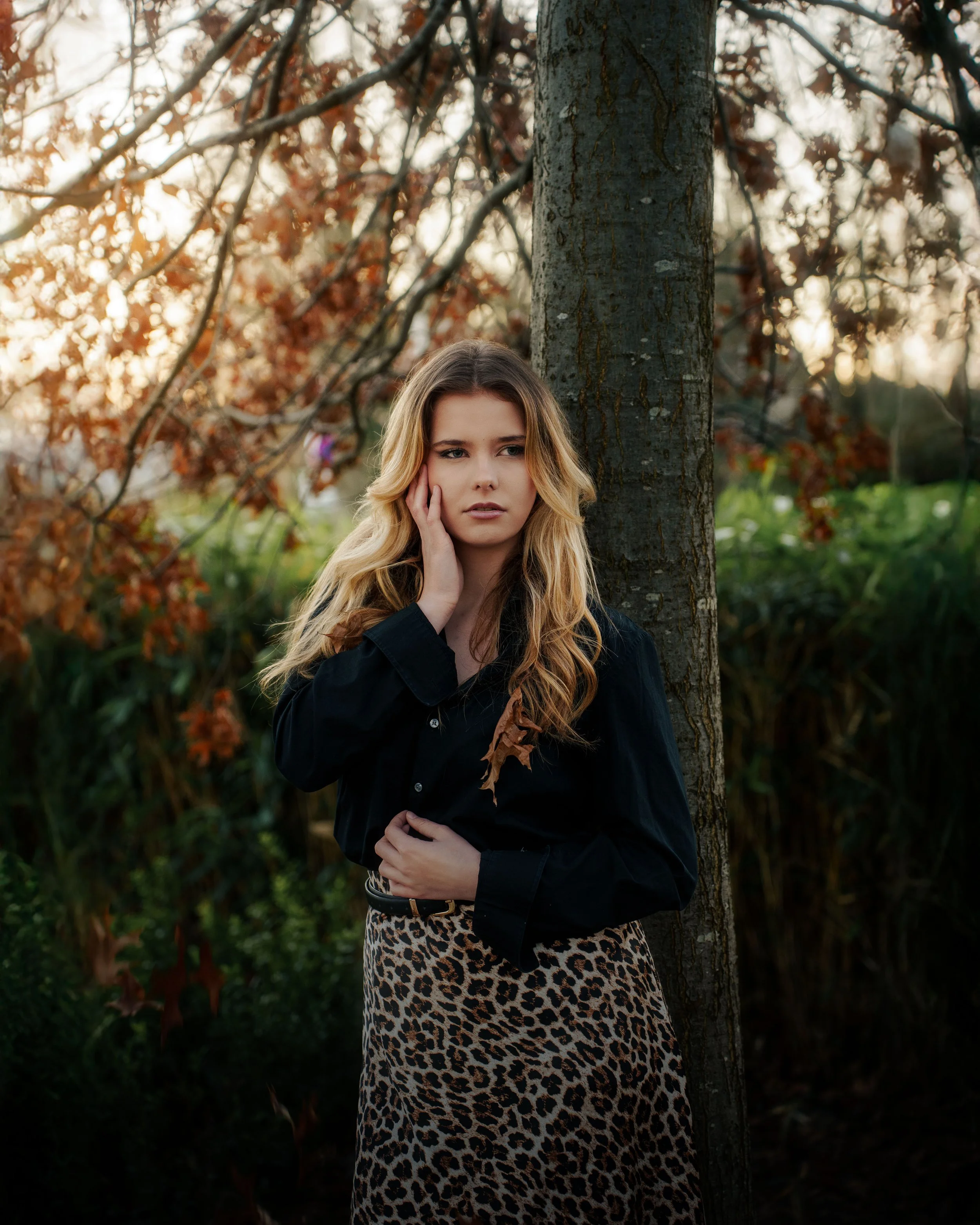 Jeune femme avec des cheveux blonds et longs, portant une chemise noire et une jupe léopard, debout près d'un arbre en forêt lors du coucher du soleil.