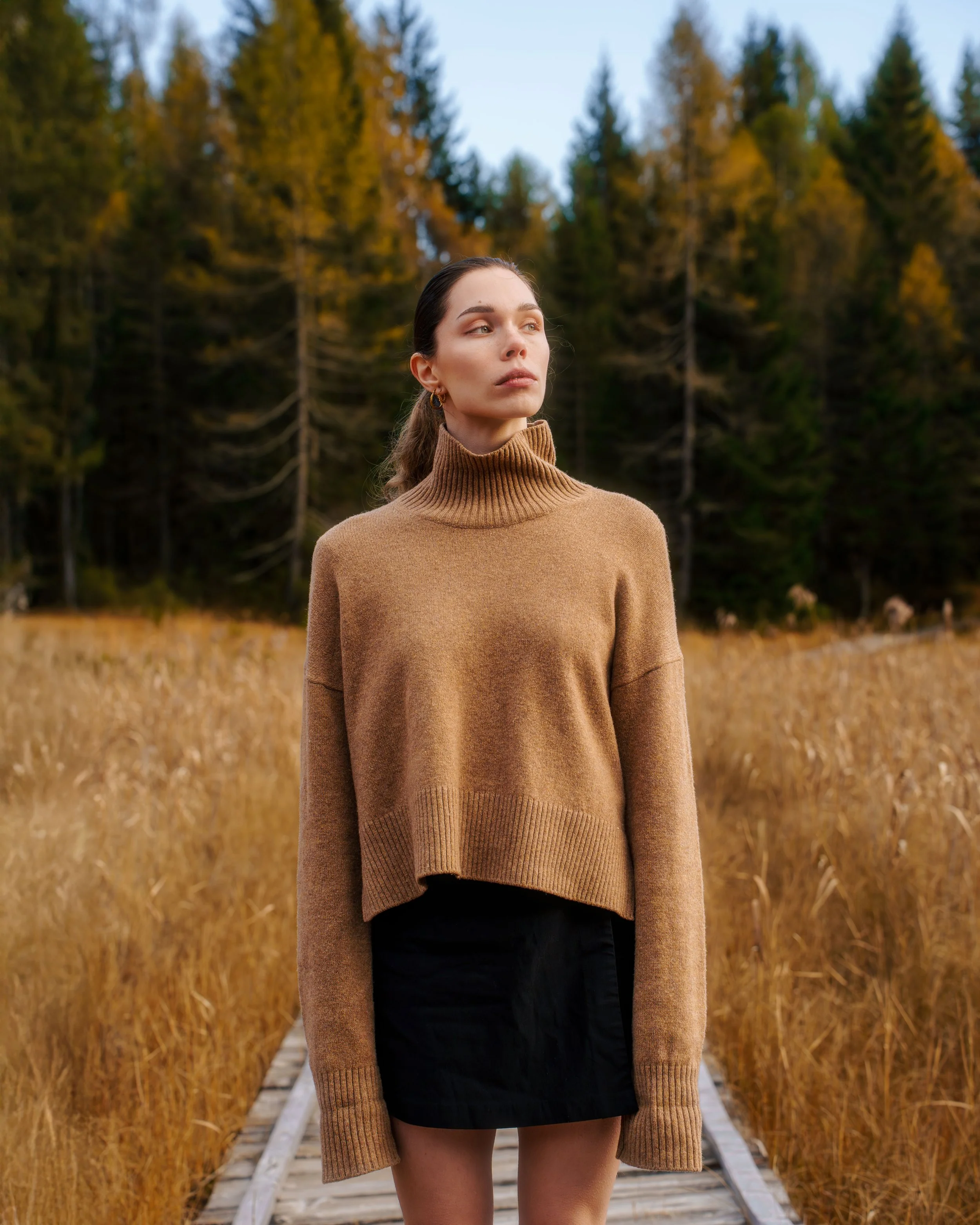 Femme debout sur une passerelle en bois dans un paysage forestier automnal, portant un pull beige à col roulé et une jupe noire.