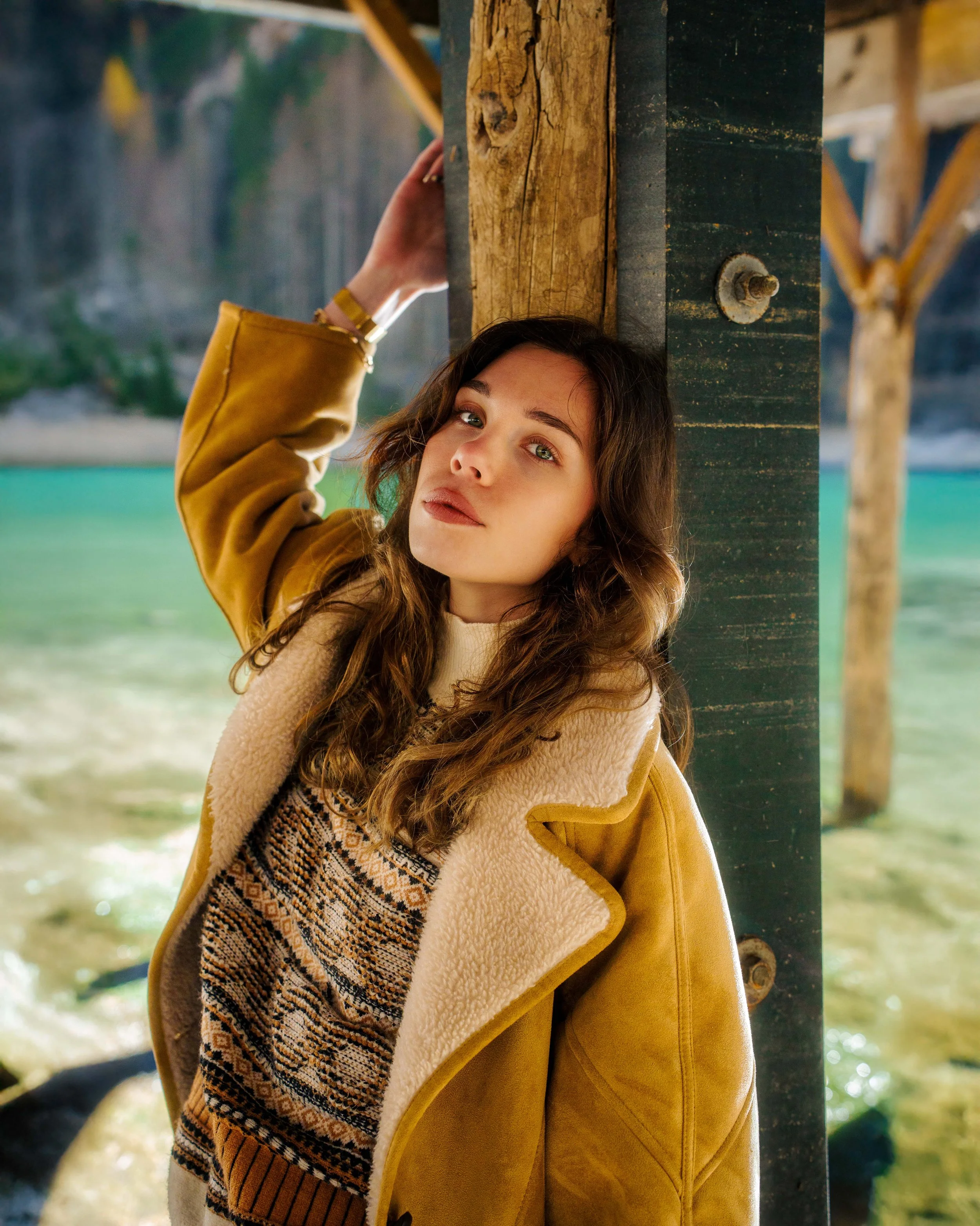 Une femme avec des cheveux bruns et peignoirs en laine, posant sous une jetée en bois avec vue sur une mer turquoise et des falaises rocheuses en arrière-plan.